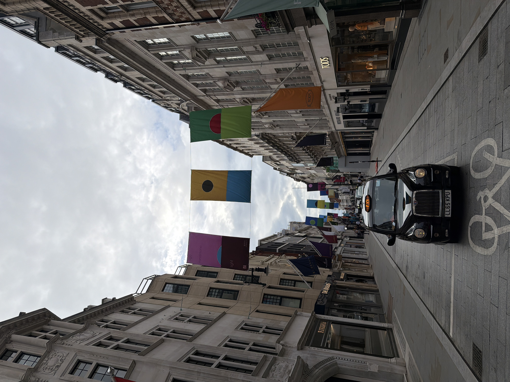 The image captures a street scene in London, United Kingdom, likely on a shopping street such as Bond Street, given the presence of luxury retail stores. In the foreground, a black London taxi is parked in a designated bay, which is marked with a white bicycle symbol on the cobblestone pavement. The taxi has its license plate clearly visible, reading "LL65 KPU".
The street is lined on both sides with tall, ornate buildings, indicative of a historic urban environment. Many of these buildings house high-end boutiques and stores, with prominent signage for brands such as "DE BEERS," "RICHARD MILLE," and "TOD'S." The facades of the buildings are a mix of stone and brick, featuring numerous windows and architectural details.
Above the street, colorful banners are strung across, adding a festive or decorative element to the scene. These banners are rectangular and appear to feature abstract designs or patterns, with some displaying text. Several flags are also flying from the buildings, including a dark blue flag with what appears to be a crest.
The sky is overcast with a muted, cloudy light, suggesting it may be a cloudy day. A few people are visible in the background, walking along the pavement and entering or exiting shops, but they are small and indistinct. The overall impression is one of a busy, upscale shopping district in London.