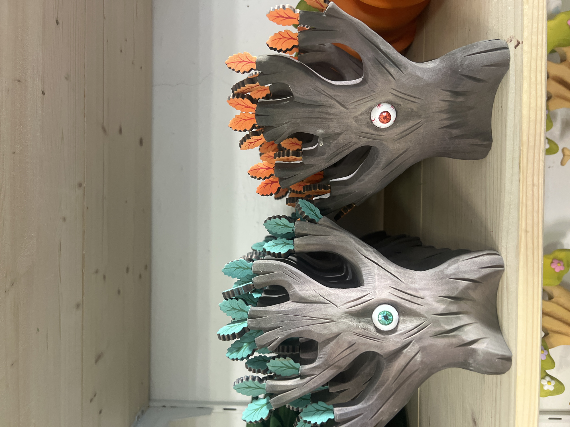 This media file shows two carved wooden, tree-like objects with cartoonish eyes. They are arranged one above the other, with the top object slightly overlapping the bottom one. Both objects are carved to resemble tree stumps with faces. The top object has orange leaf-like decorations and a red and white eye. The bottom object has green leaf-like decorations and a green and white eye. The objects are set against a background of light-colored wooden planks on the left and a white wall on the right. There are hints of other decorative objects to the right, including what appears to be a carved pumpkin and a wooden cutout with flowers. The lighting suggests it is daytime, and there are no obvious indicators of weather or a specific location, other than the interior setting. The overall tone is whimsical and decorative. There is no visible text in the media file.