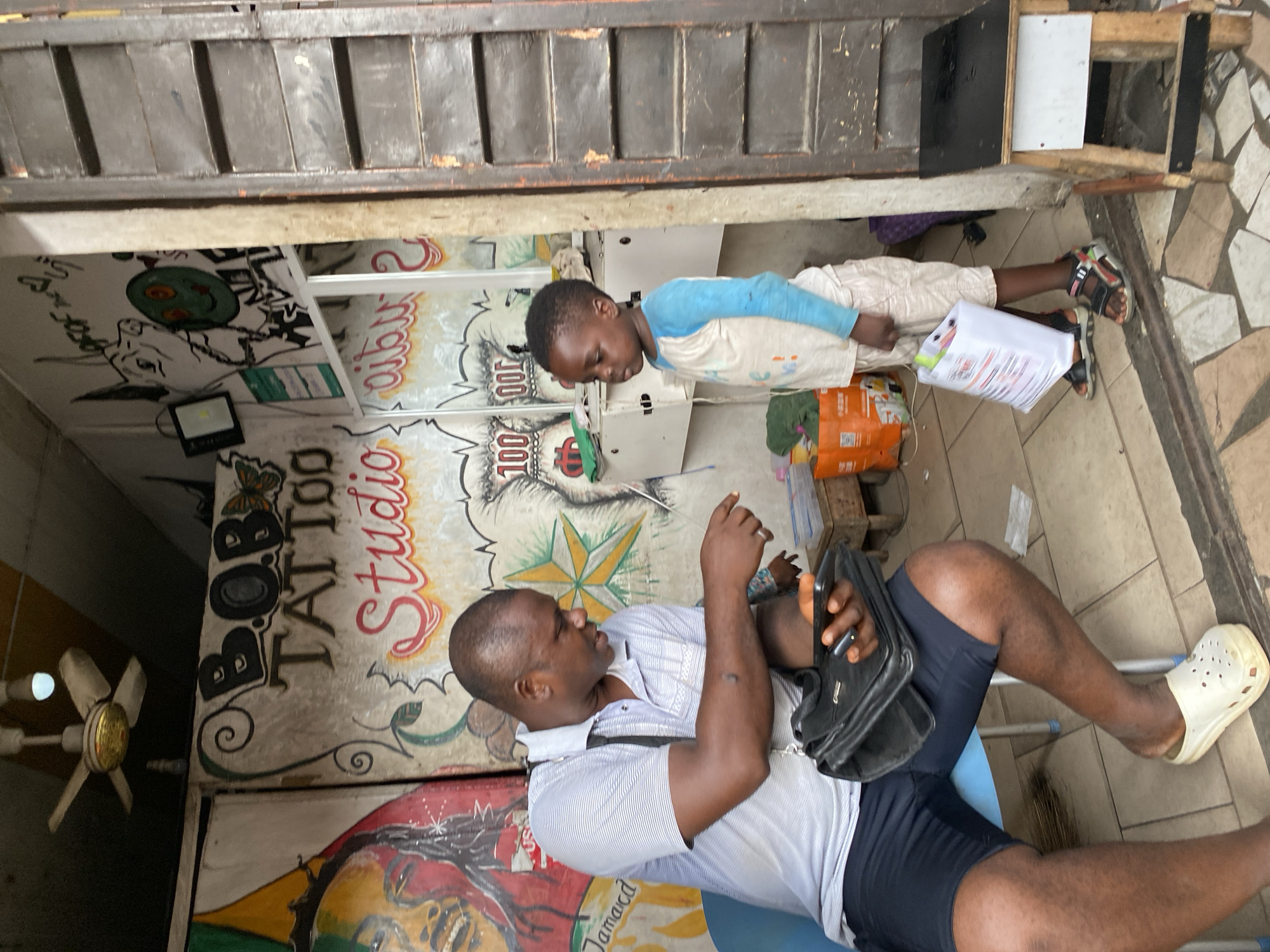 The image shows an outdoor scene in Lekki, Nigeria, in front of a "BOB Tattoo Studio". A man in a light blue striped shirt and dark blue shorts sits on a light blue plastic chair, holding a black shoulder bag and looking towards a young boy in a blue-sleeved shirt and light-colored pants. The boy is standing and holding a white paper or document. A fan is visible on the left side of the image. The studio has colorful artwork on its exterior.