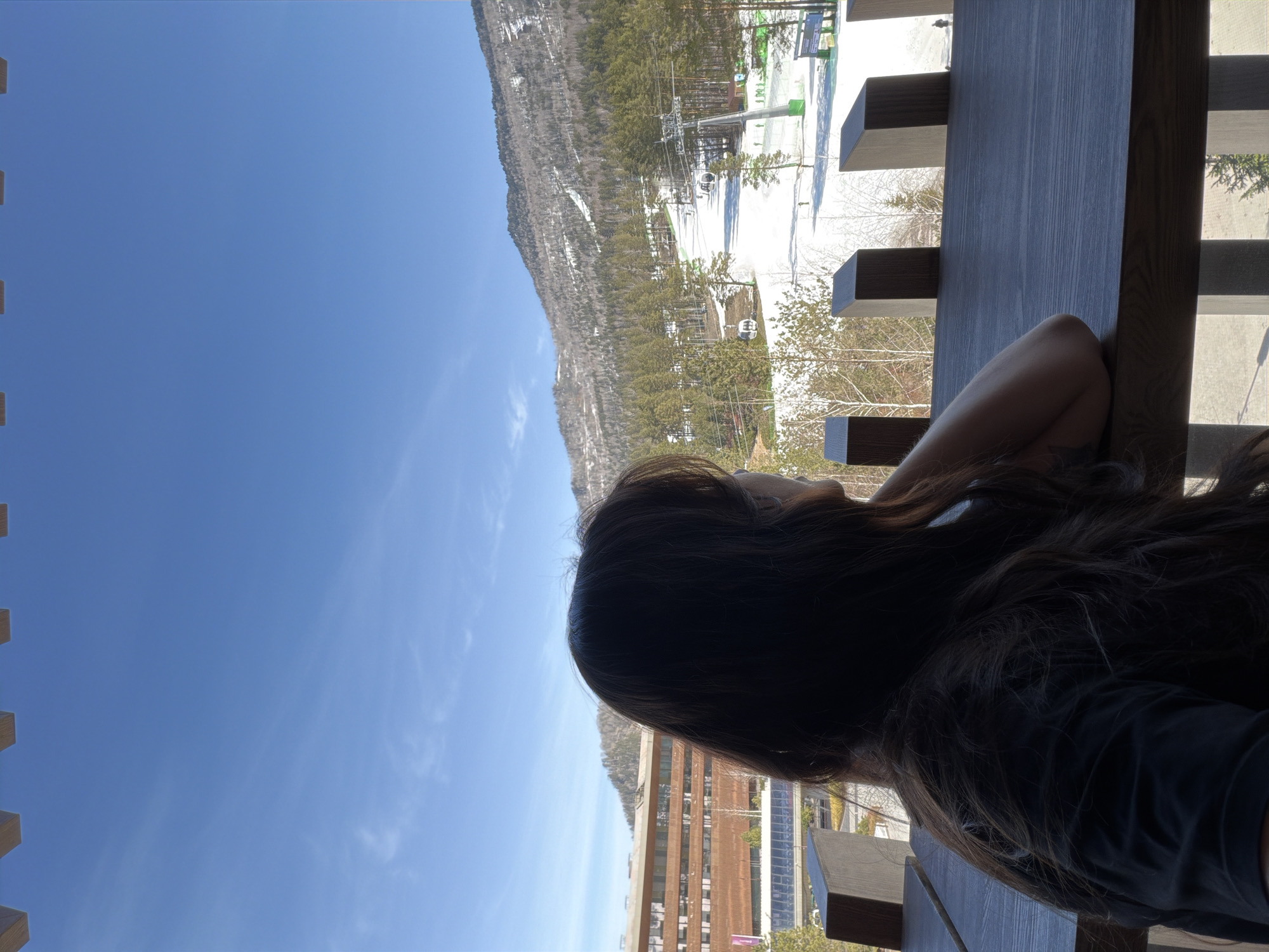 A woman sits at a wooden structure overlooking a snowy mountain and a building. The sky is blue. It is not possible to confirm the location of the image based on the visual information.