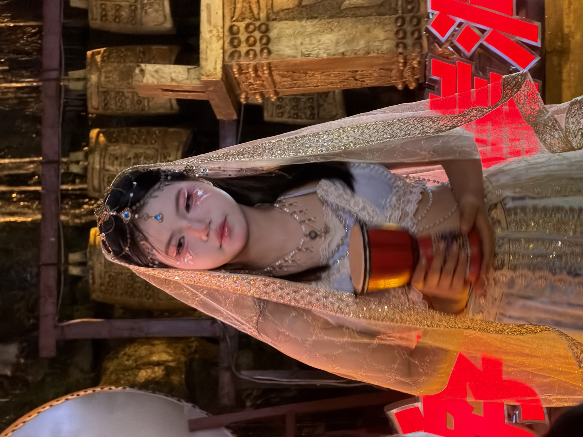 A young woman in a light blue and gold ornate outfit, complete with a veil and elaborate headpiece, stands holding a red drum. Her makeup features bright pink eyeshadow and glitter accents on her cheeks. The background consists of a display of large, ornate bells, suggesting a cultural or historical setting. To the right, a tall, gilded structure with decorative elements is visible. Large, red Chinese characters, partially obscured, are present in the foreground on both sides of the woman. The lighting is dim, with warm tones illuminating the scene, particularly the woman and the bells.