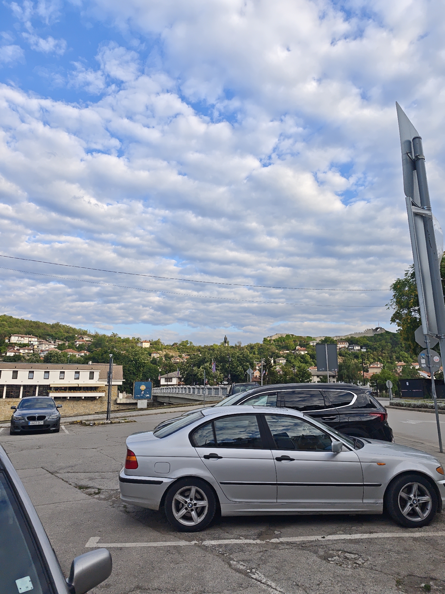 A silver BMW sedan is parked in a lot with a black SUV behind it. In the background, a bridge spans a body of water, and a hillside town with red-tiled roofs and lush greenery is visible. A fortress or castle wall crowns the hill. The sky is filled with white, fluffy clouds against a blue backdrop. The scene appears to be in Lovech, Bulgaria, given the distinctive architecture and the fortress on the hill, which strongly resembles the Hisarya Fortress.