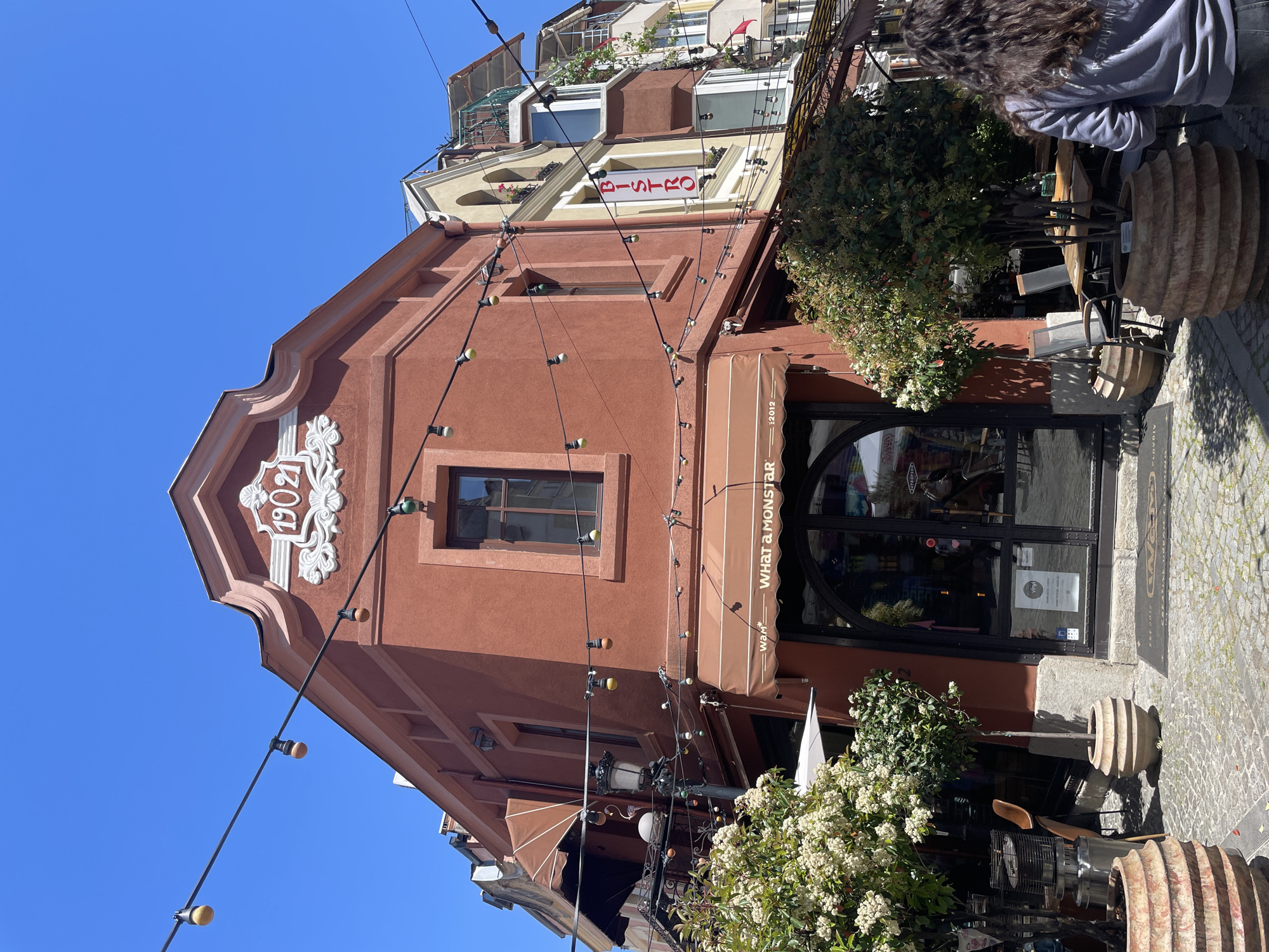 The image shows the façade of a reddish-brown building in Plovdiv, Bulgaria. Above the arched doorway, a brown awning reads "WHAT a MONSTAR". Above the awning is a rectangular window. Higher up, near the roofline, a decorative white emblem displays the number "1902". Strings of lights hang across the building's front. Further up, a building with a "BISTRO" sign is visible. A person with curly brown hair partially blocks the upper right corner of the image. Outside the building is a stone-paved area. Decorative planters made of light brown stacked rings stand near a table and chairs. Flowering bushes are planted near the entrance. The sky is clear and blue.