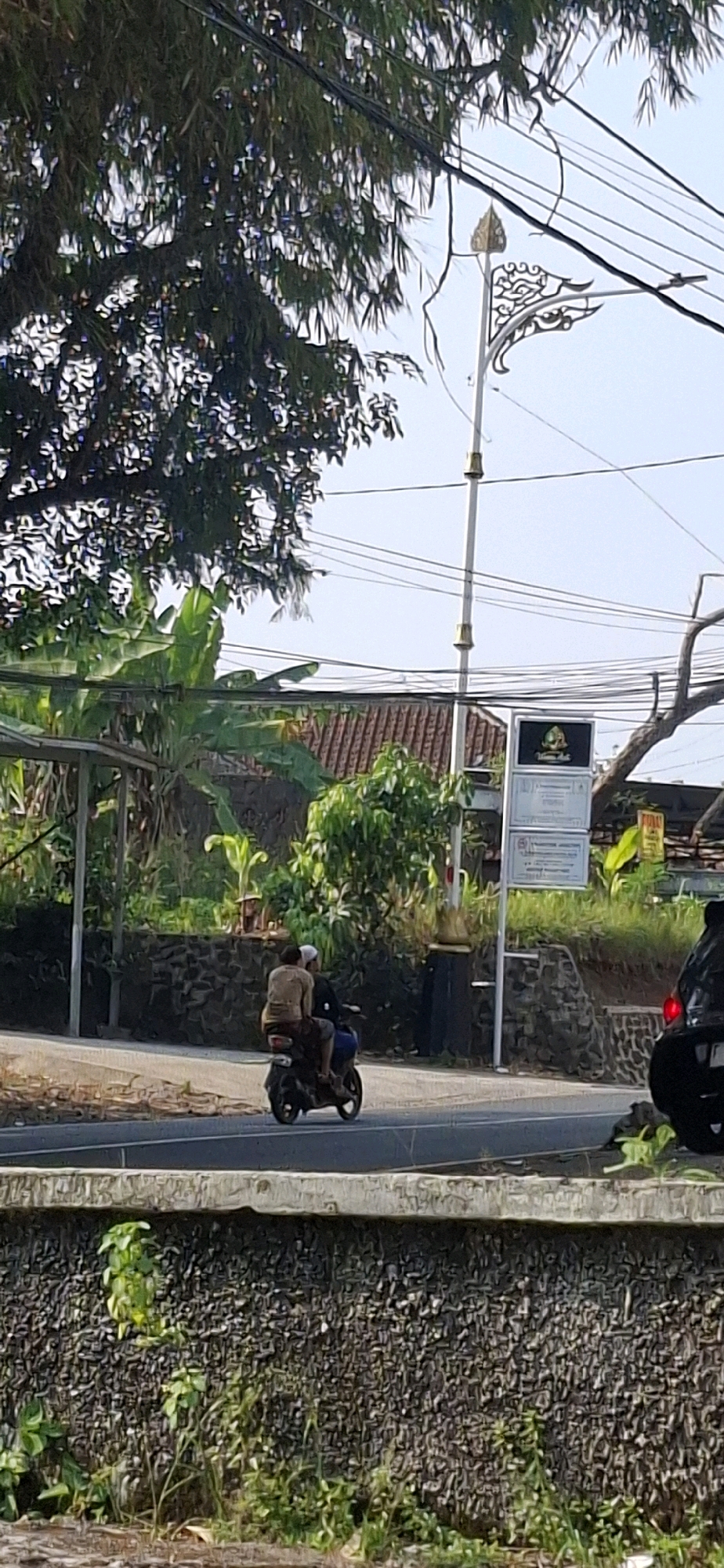 In Cilaku, Indonesia, two individuals ride a motorcycle down a paved road. The passenger, positioned behind the driver, is wearing a light brown shirt and dark pants, with their head covered by a white cloth. The driver is wearing a dark blue top. The motorcycle is moving from left to right. Behind them, a stone wall runs along the side of the road, topped with vegetation. In the background, there are trees, a building with a tiled roof, and a decorative street lamp with ornate metalwork. Electrical wires are strung across the sky. A signpost with multiple white panels and a dark green logo is visible to the right of the street lamp. Further to the right, the rear of a black car is partially visible. The scene appears to be set during daylight.