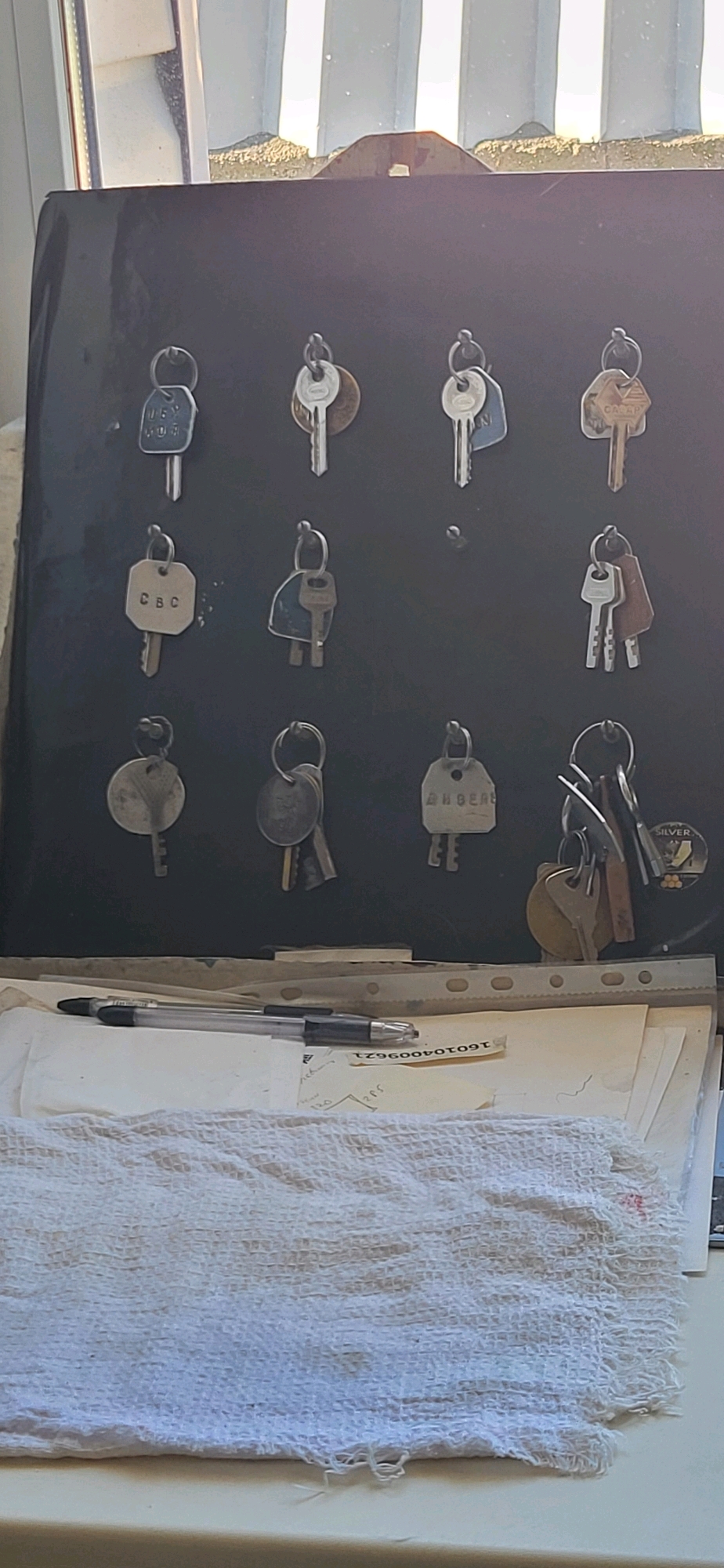 The image depicts a collection of old keys hanging on a dark-colored board, possibly a key organizer. The board is mounted against a wall and is situated near a window that allows natural light to enter the scene. The keys vary in shape, size, and material, with some appearing to have tags or engravings. Below the key organizer, there is a stack of papers and a pen, suggesting a workspace or a place where documents are stored. A white, textured cloth lies in the foreground, partially covering the papers. The overall scene suggests a domestic or office environment. The image does not provide any specific visual cues to locate it in the city of Oskemen, Kazakhstan.