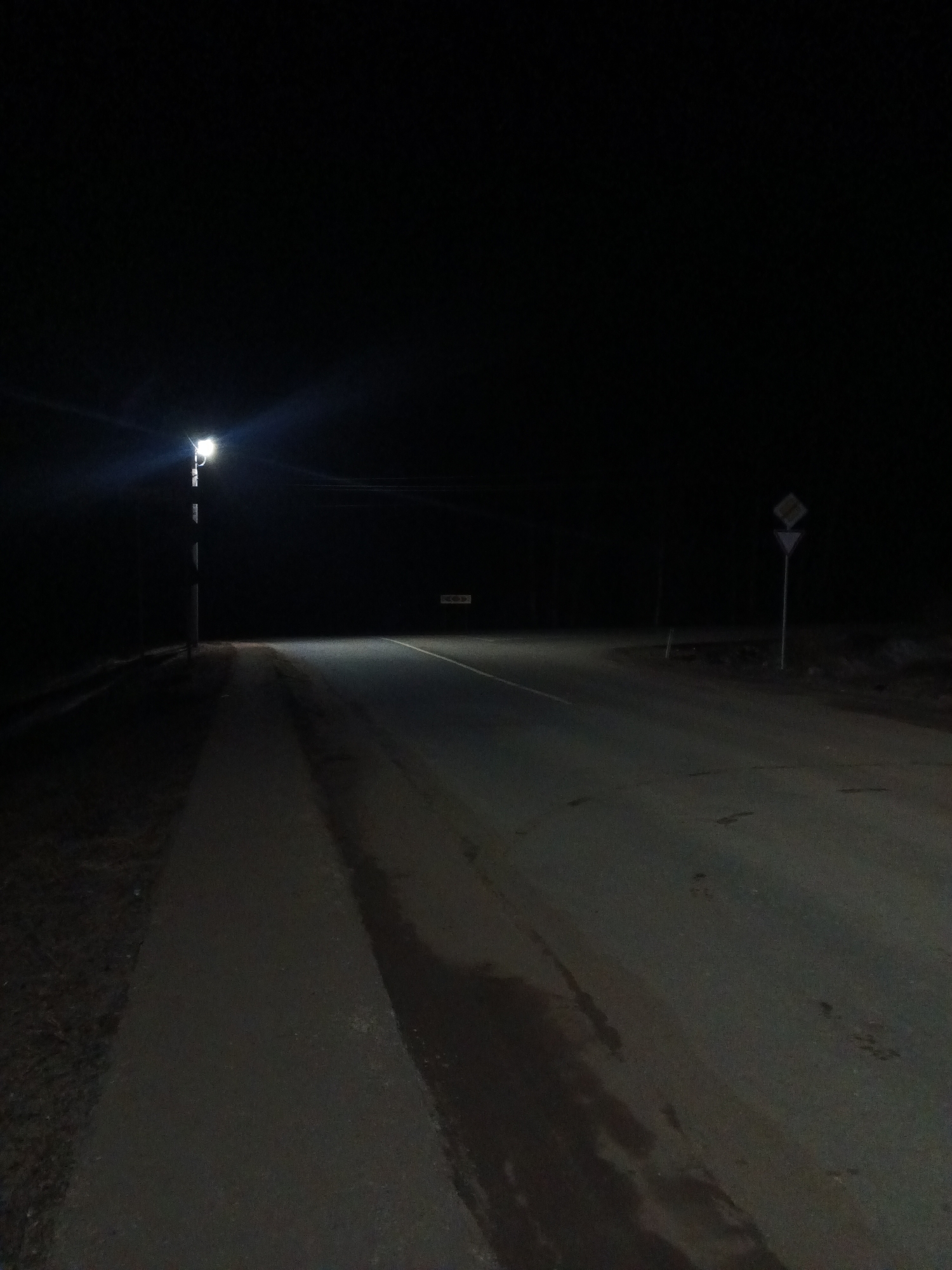 The image depicts a dark, seemingly deserted street in Rusalevka, Russia at night. A single street lamp illuminates a section of the road and a paved walkway. Further down the road, a sign is barely visible in the distance. To the right of the road, a road sign is visible. The road surface shows signs of wear and possible dampness. The surrounding environment is obscured by darkness. There are no visible people or subjects in the image. The scene suggests a quiet and isolated location within the city.