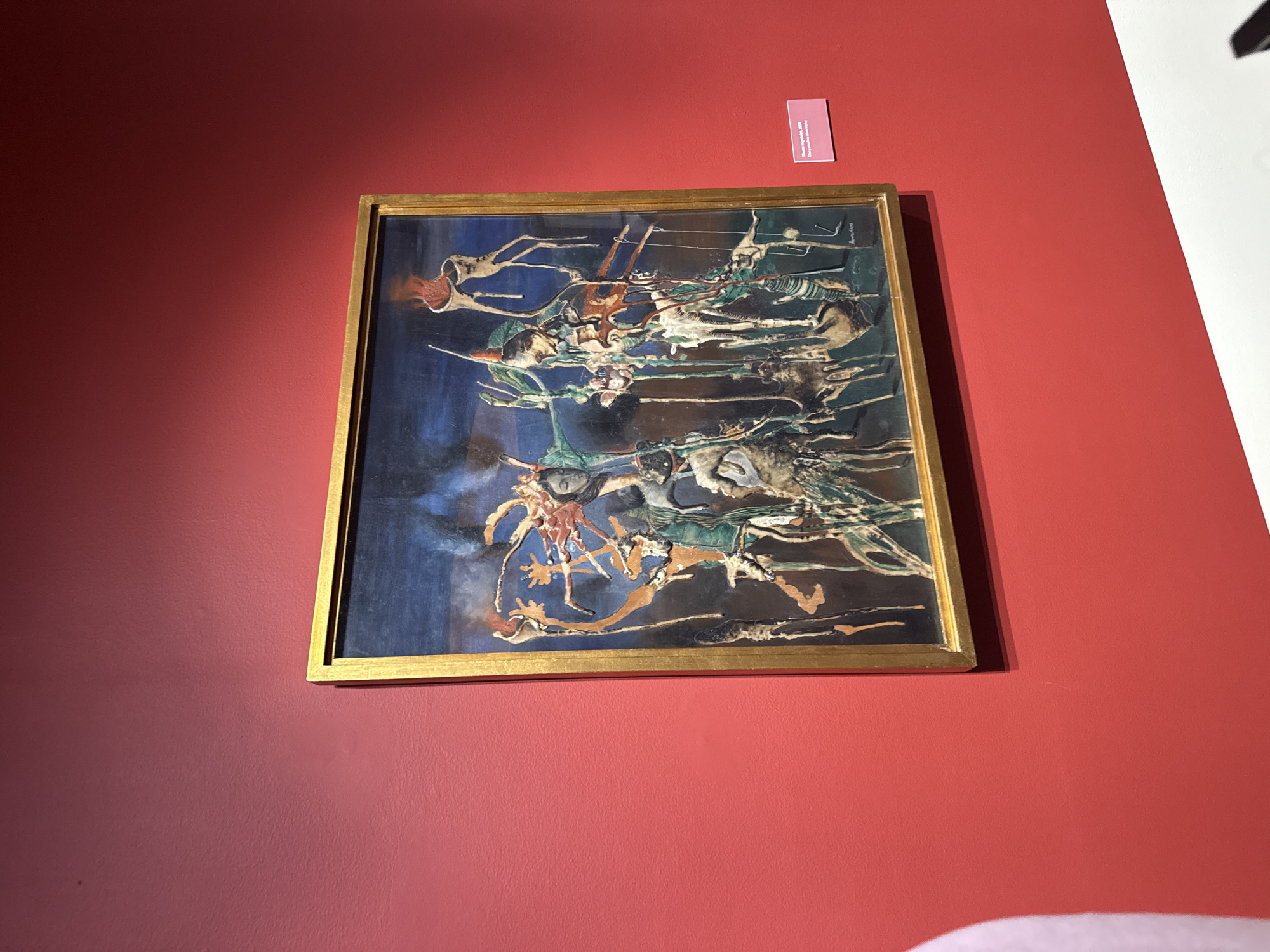 A framed painting hangs on a red wall at an art gallery in Mexico City, Mexico. The painting depicts abstract figures in an impressionistic style, featuring a gold frame and a small information placard above it on the wall. The wall is well-lit, with a corner of a white wall visible on the upper right.