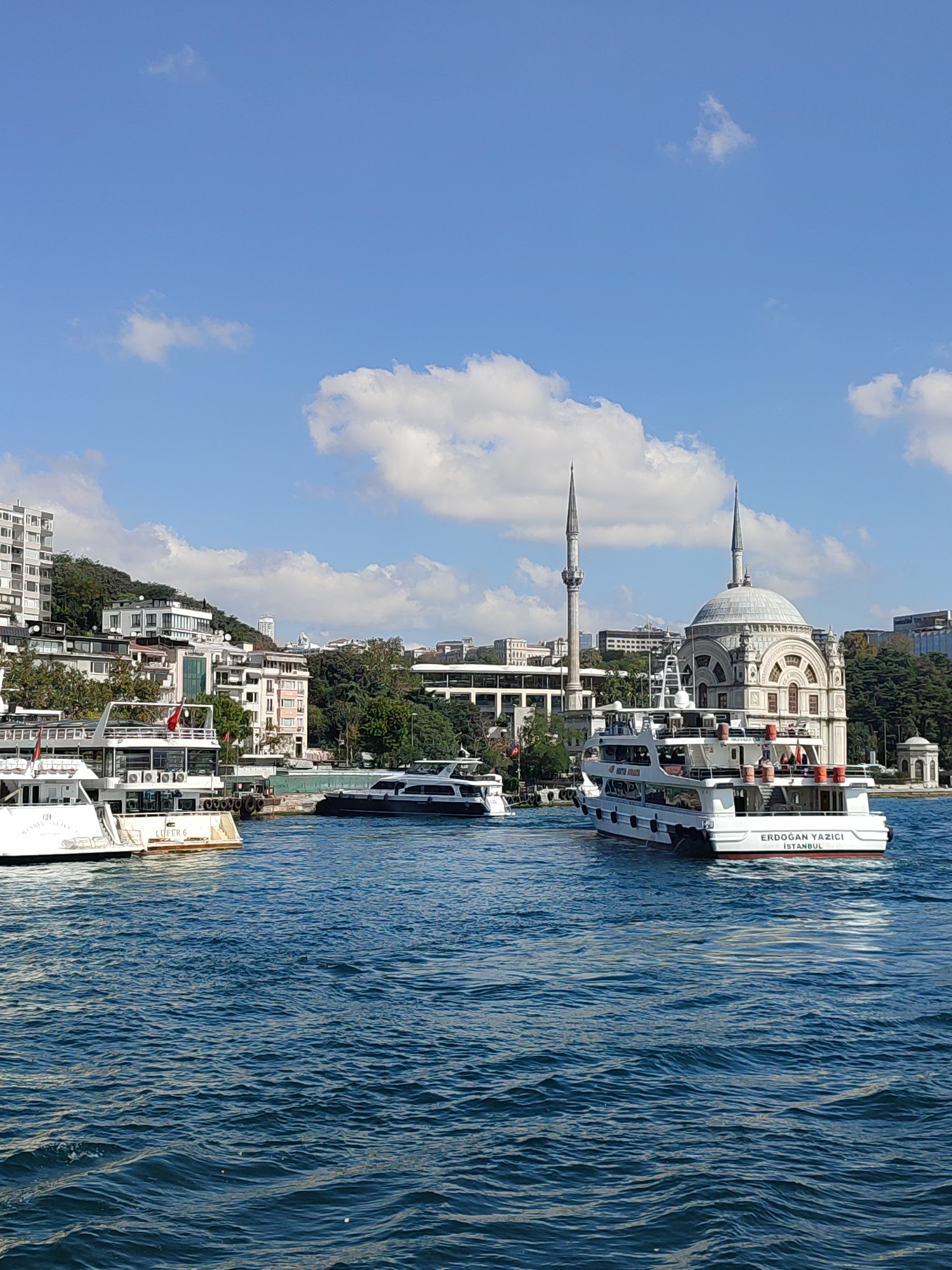 A waterfront scene captures a bright, sunny day with a clear blue sky dotted with fluffy white clouds. The Bosphorus strait is filled with deep blue, choppy water, reflecting the daylight. In the foreground, several boats are visible. The most prominent is a white ferryboat with multiple decks, bearing the inscription "ERDOĞAN YAZICI ISTANBUL" on its stern. Another white ferry, partially obscured, is docked to the left.
In the middle ground, the Istanbul skyline rises. A distinctive mosque with a large dome and two tall minarets stands out. Behind the mosque, a cluster of apartment buildings and other structures are nestled on a tree-covered hillside. The architecture suggests a blend of modern and traditional styles. The overall atmosphere is tranquil and picturesque, depicting a vibrant urban landscape by the water.