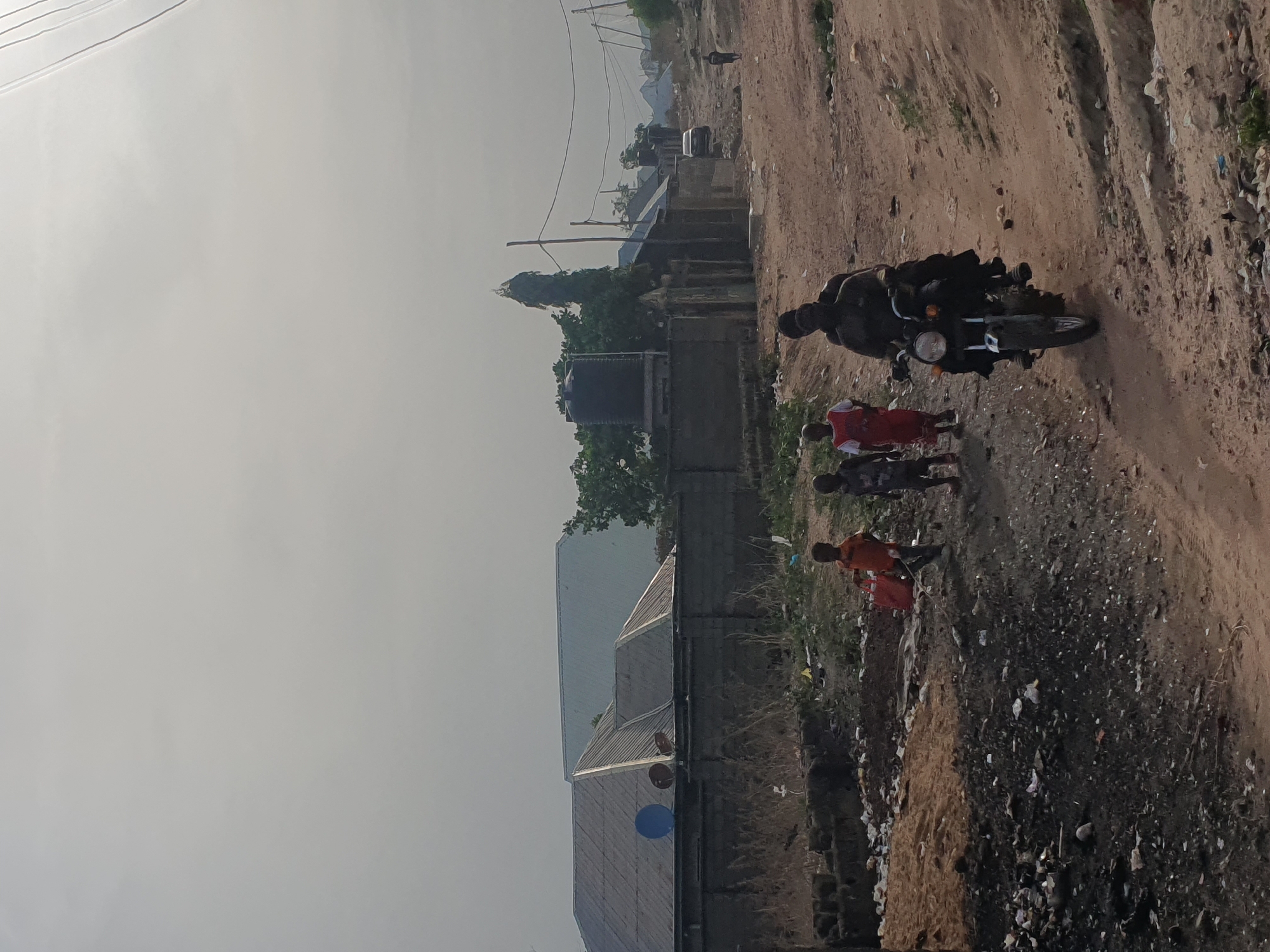 The image captures a semi-rural or peri-urban scene in Lokoja, Nigeria, under an overcast sky or during a soft-light period like early morning or late afternoon. A wide, unpaved dirt path runs along the right side of the frame, showing signs of vehicle tracks and scattered debris.
On the path, a person dressed in dark clothing is riding a dark-colored motorcycle, facing away from the viewer. Behind the motorcycle, three children are walking; two are wearing red or orange shirts, and one has a darker top.
To the left of the dirt path, several modest buildings with corrugated metal roofs are visible. Some sparse vegetation and trees are interspersed among the structures. A black water tank on a stand is prominent near one of the buildings. In the background, a tall, slender structure, possibly a minaret or a water tower, stands out above the trees and distant buildings. Overhead, power lines or communication cables are visible across the hazy sky. The ground appears dry and dusty. No discernible text is visible in the image. The overall scene depicts a slice of daily life and infrastructure in the area.