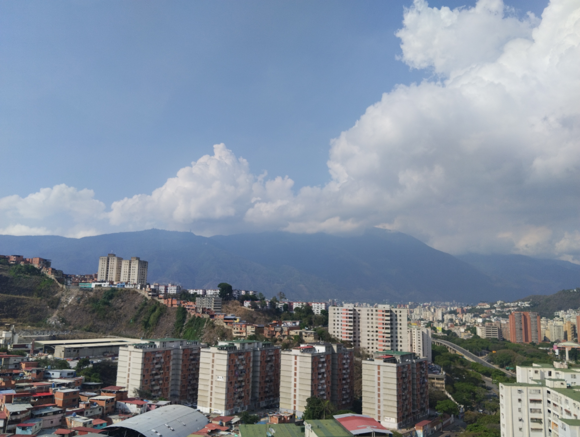 This is a high-angle, wide shot of a dense cityscape, likely Caracas, Venezuela, as indicated by the context. The scene is dominated by urban development spread across hilly terrain, with imposing mountains forming the backdrop under a partly cloudy sky.
In the foreground and mid-ground, a mix of residential buildings is visible. There are numerous multi-story apartment blocks, some with uniform facades, others showing exposed brick or concrete. Many smaller, less formal structures with colorful roofs are clustered together on the hillsides, characteristic of informal settlements or barrios often found in Latin American cities. Roads and a highway segment wind through the urban landscape, partially obscured by trees and other buildings. Some industrial-looking structures with large roofs are also present.
The background features a grand mountain range, appearing somewhat hazy or covered by light clouds, stretching across the horizon. The sky above is a clear, bright blue with scattered white and light grey cumulus clouds, indicating fair weather. The sun is shining brightly, casting distinct shadows and suggesting it's late morning or early afternoon. No people or specific activities are discernible, and no text is visible within the image.