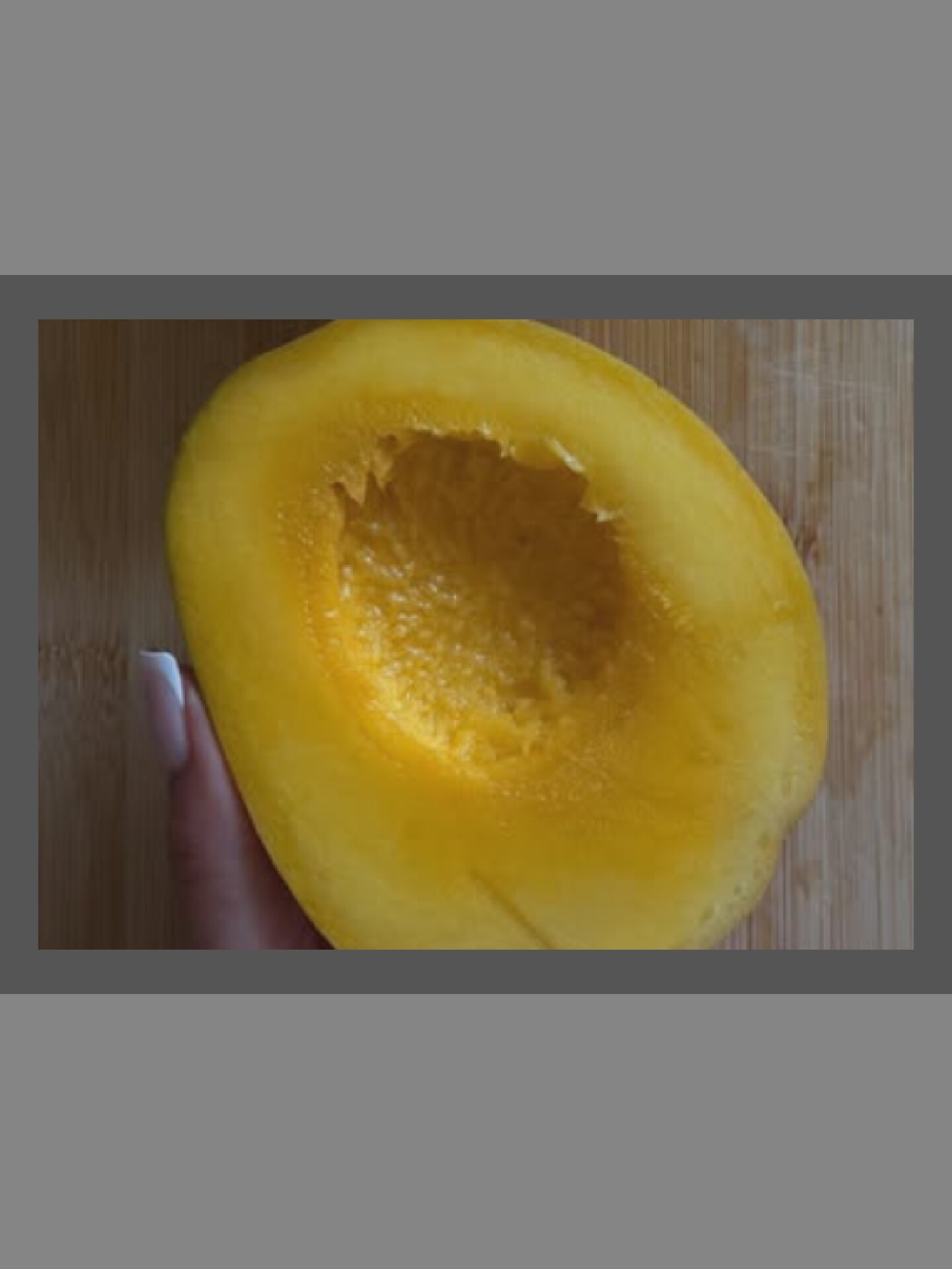 An overhead shot shows a halved yellow spaghetti squash on a wooden cutting board. A person's hand with manicured nails is holding the squash from the left side of the frame. The squash is bright yellow, with a textured interior and a smooth exterior. The cutting board has a light brown wood grain pattern. The image is cropped with gray bars at the top and bottom.