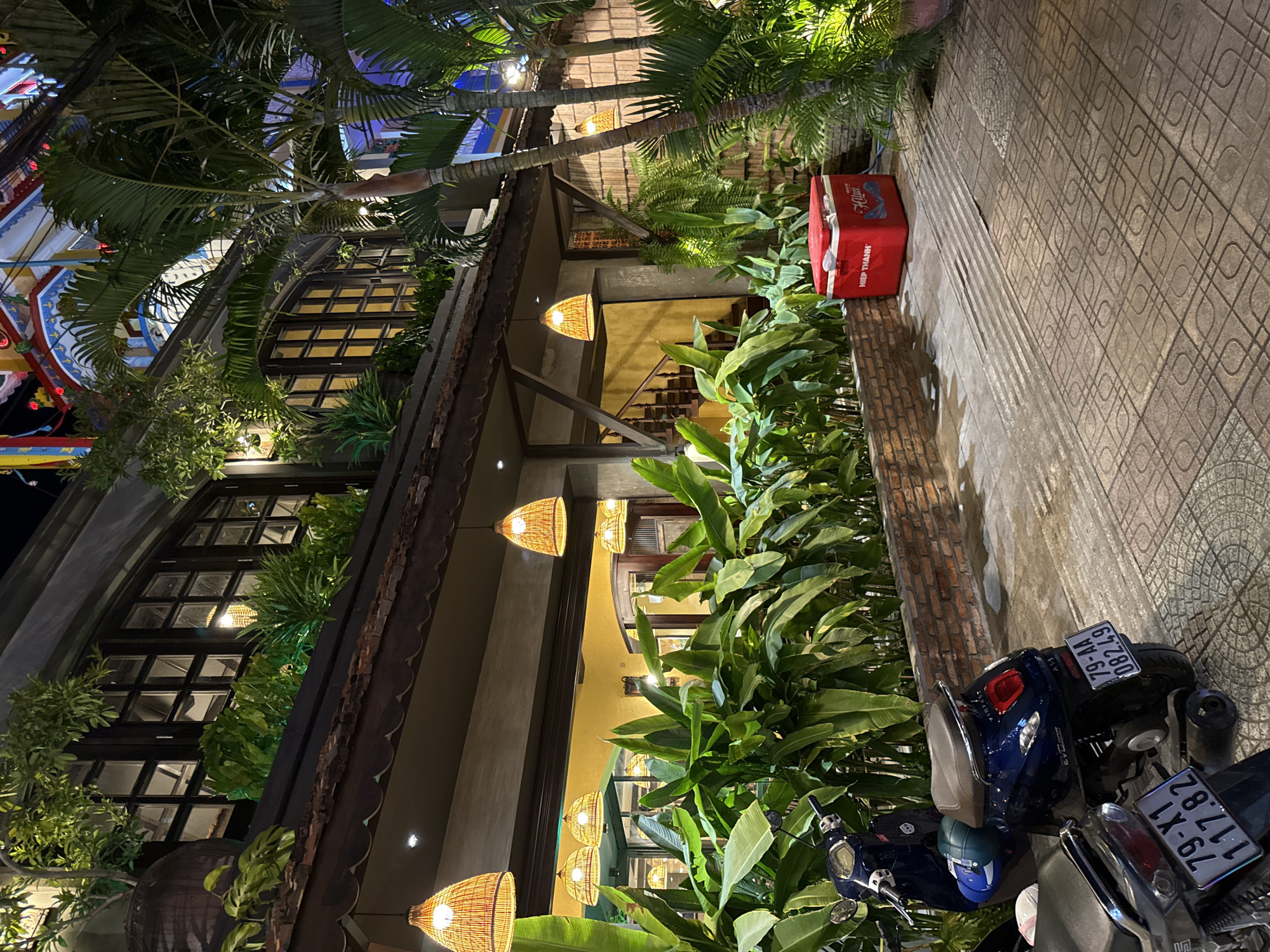 The image shows a nighttime street scene in Nha Trang, Vietnam. In the foreground, two dark blue scooters are parked on a tiled sidewalk. The scooter on the right has a license plate that reads "79-AA 082.49". The scooter on the left has a license plate that reads "79-X1 117.82".
Behind the scooters is a wall made of red bricks. Beyond the wall, a building with a second floor is visible. This building has large windows with dark frames and appears to be illuminated from within. Several hanging pendant lights with woven shades hang from the underside of an overhang in front of the building. Lush green plants, including large-leafed tropical plants and palm trees, surround the building and contribute to a vibrant, somewhat tropical atmosphere.
To the right of the scooters and plants, a bright red cooler box is visible on the ground. It has the words "HIỆP THÀNH" printed on it in white letters, along with a logo that appears to be for a beverage company.
The overall impression is a well-lit, lively commercial or residential area in Nha Trang at night, characterized by its architecture, abundant greenery, and the presence of motorbikes, which are a common mode of transport in Vietnam.