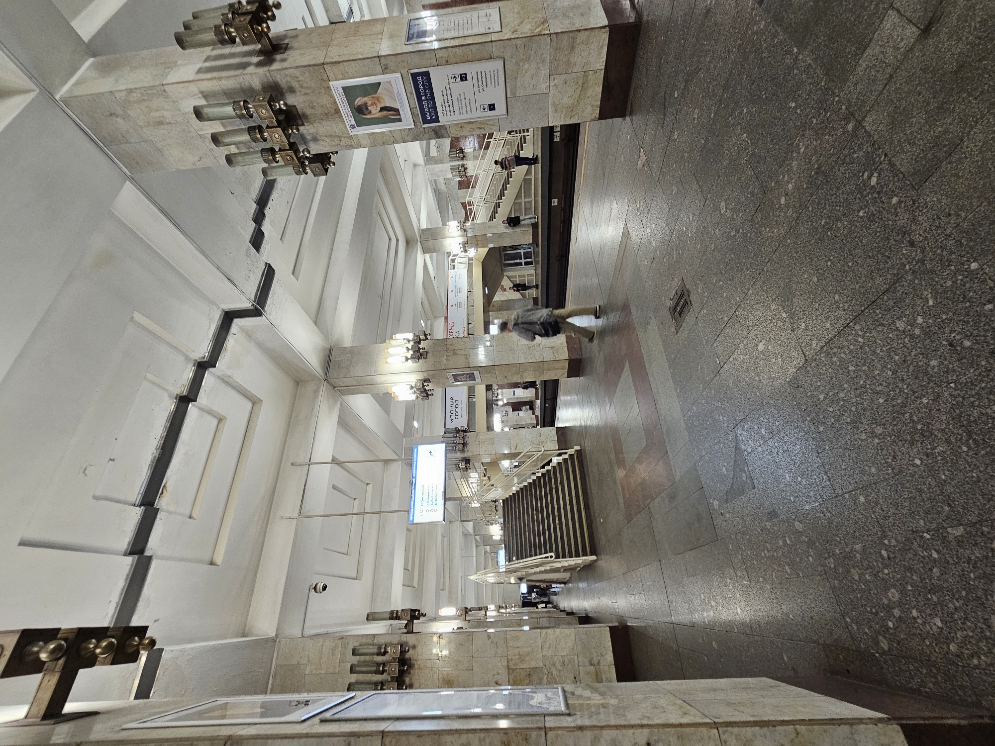 This image depicts the interior of a metro station, likely in Nizhny Novgorod, Russia, given the location context. The scene is characterized by its utilitarian architecture, featuring marble-clad pillars and walls, a tiled floor, and a high, vaulted ceiling with embedded lighting fixtures.
Several people are visible in the station, navigating the space. One person, walking away from the camera, is dressed in a grey jacket and dark pants, carrying a light-colored bag. Further in the background, other individuals can be seen on a staircase and near a metro platform. The station appears to be moderately busy, with individuals moving about.
Notable details include the prominent use of marble and granite, indicative of Soviet-era metro station design. Signs with text are present on the pillars, including one that reads "ВЫХОД В ГОРОД" (Exit to the city) with an arrow pointing towards the right, and another sign with Cyrillic text and pictograms suggesting directions or information about the metro lines. Another sign appears to be an advertisement or information poster. The lighting suggests it is daytime, though the interior setting makes it difficult to ascertain external weather conditions. The overall atmosphere is functional and somewhat impersonal, typical of public transportation hubs. The orientation of the image is skewed, with the scene appearing to be shot from a tilted perspective.