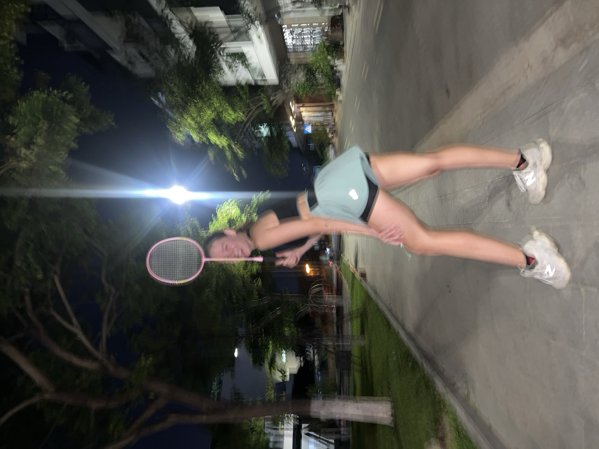 A person with fair skin, wearing a black top, light blue shorts, and white sneakers, holds a pink badminton racket. The person stands on a sidewalk bordering a grassy area and a street. Buildings and trees are visible in the background. The setting is at night with artificial lighting present. The location is possibly in Nam Nha Trang, Vietnam.