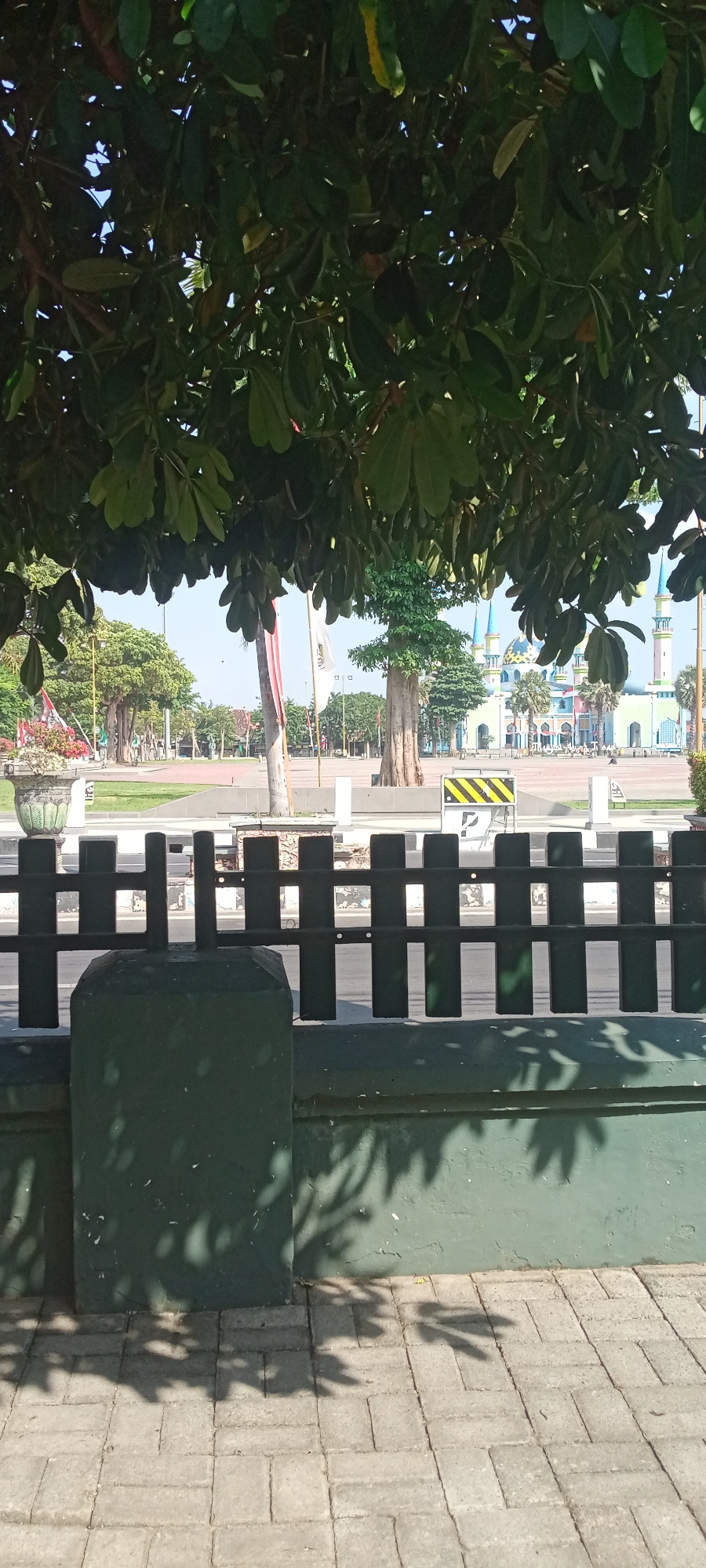 The image depicts a view from behind a dark green fence looking out onto a public square in Tuban, Indonesia. The foreground features a paved walkway with brick-like patterns and a dark green wall with visible leaf shadows. Beyond the fence, a road runs horizontally, bordered by a white fence. In the midground, a grassy area is visible with trees and a decorative pot containing red and white flowers. Further in the background, a large, light blue mosque with colorful domes and minarets stands prominently. The sky is clear and blue. There are several people walking in the distance near the mosque and in the open square, though they are too small to discern details. Two flags, one red and white and another white, are planted on poles near a tall, thin tree. A yellow and black striped warning sign is also visible on the road. The overall scene suggests a bright, sunny day in a public space adjacent to a significant religious building.