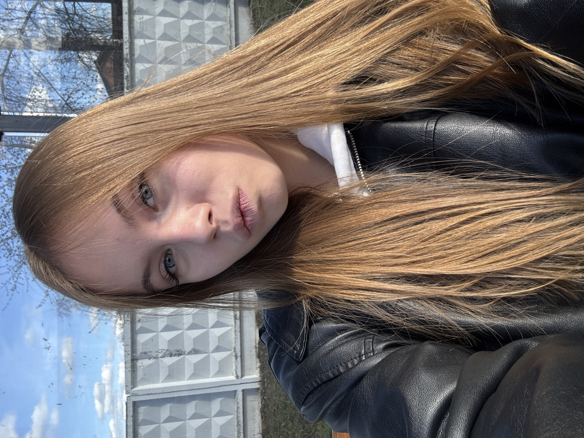 A young woman with long, light brown hair is the primary subject of this photograph. She is wearing a black leather jacket and a white hooded top. The background includes a gray concrete fence with a geometric pattern. Beyond the fence, a blue sky with tree branches is visible. The location is identified as Chaikovsky, Russia. The image is taken at a close range, focusing on the woman's face.