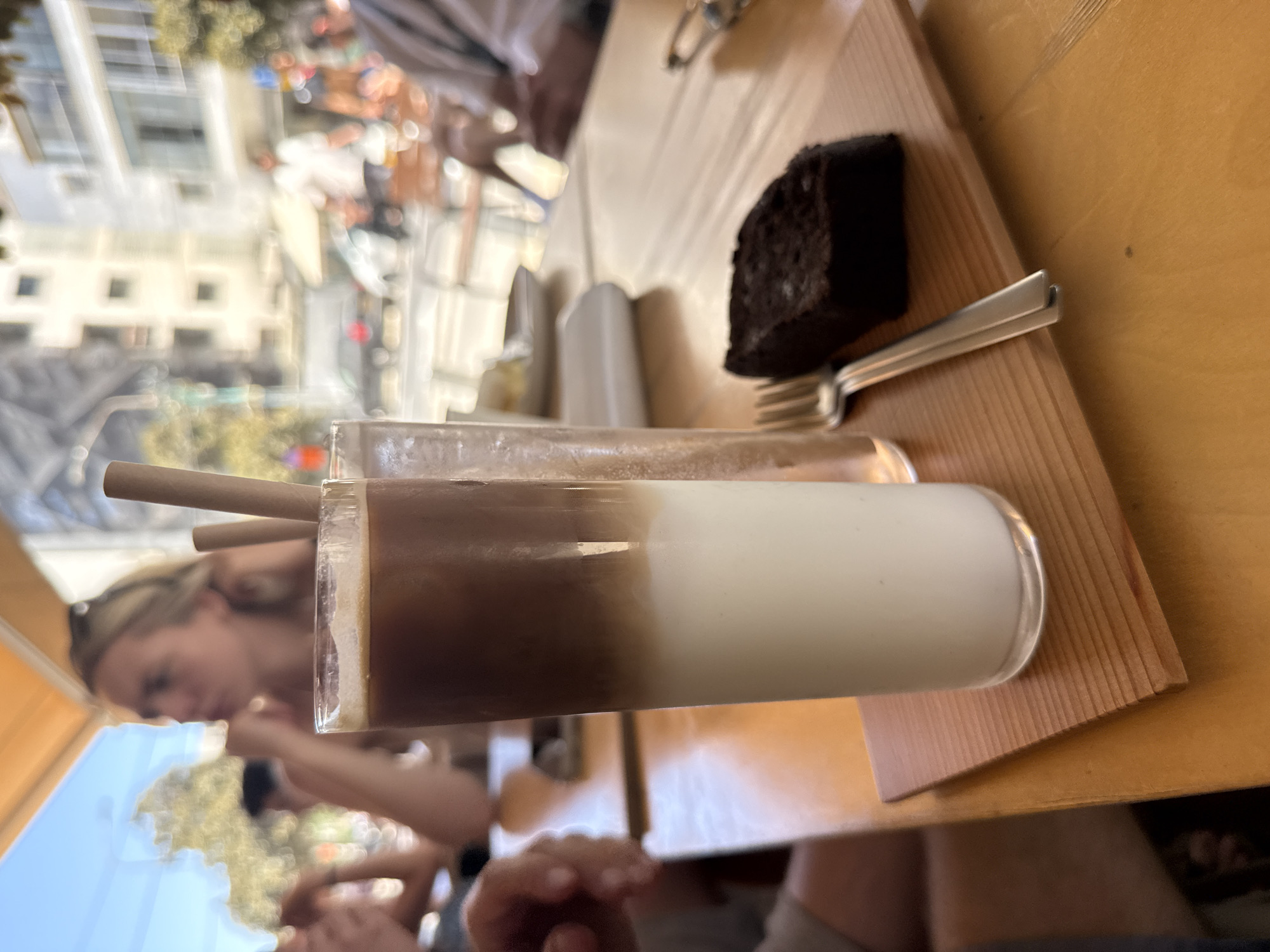 A tall, clear glass containing a layered iced coffee, consisting of a white lower layer and a dark brown upper layer with minimal froth, is centrally positioned on a light brown wooden slatted coaster. Adjacent to the coffee glass, a rectangular slice of dark cake and a silver fork are also resting on the coaster. To the right of the coffee, a second clear glass containing liquid and two light brown straws is partially visible. These items are placed on a wooden table with a light orange-brown finish. In the blurred left foreground, a person's hand and arm are visible on the table. In the out-of-focus midground, a blonde woman with sunglasses on her head is depicted with her chin resting on her hands, looking to the left. Other indistinct figures are present in the background, suggesting a social setting. The scene is an outdoor cafe situated under a light orange awning. The background reveals an urban environment consistent with the city of Tel-Aviv, Israel, showcasing multi-story residential and commercial buildings, parked and moving vehicles, and pedestrians on a street under bright daylight conditions.