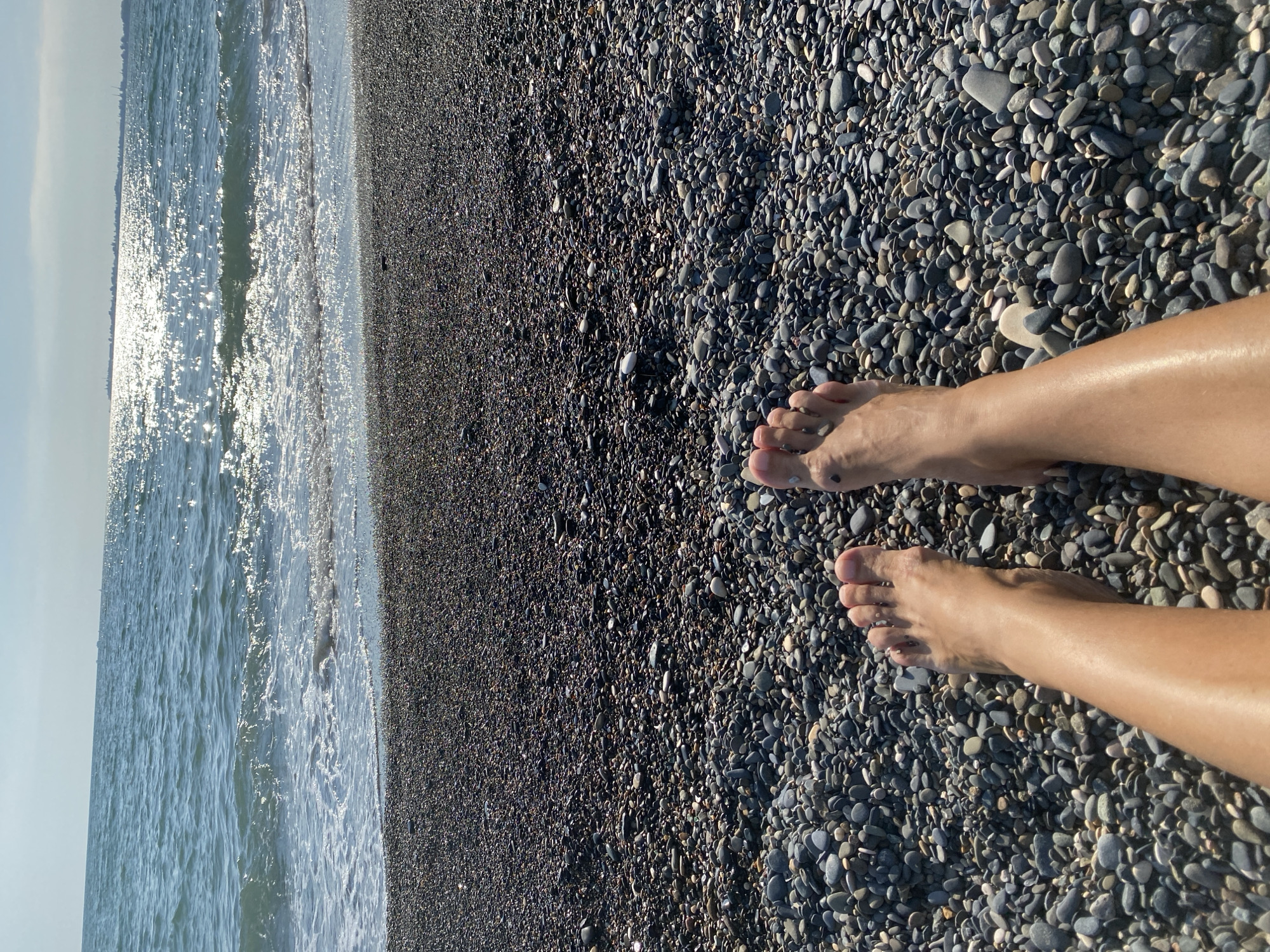 A first-person perspective shows a pair of human legs and feet resting on a dark, pebble-strewn beach. The pebbles are varied in size and color, ranging from dark gray to lighter brown and white. The ocean stretches out in the background, with gentle waves lapping at the shore. Sunlight glints off the surface of the water, creating a shimmering effect. The sky above is clear and bright. The scene is set in Chabkwara, Abkhazia, a location known for its pebble beaches and clear sea waters.