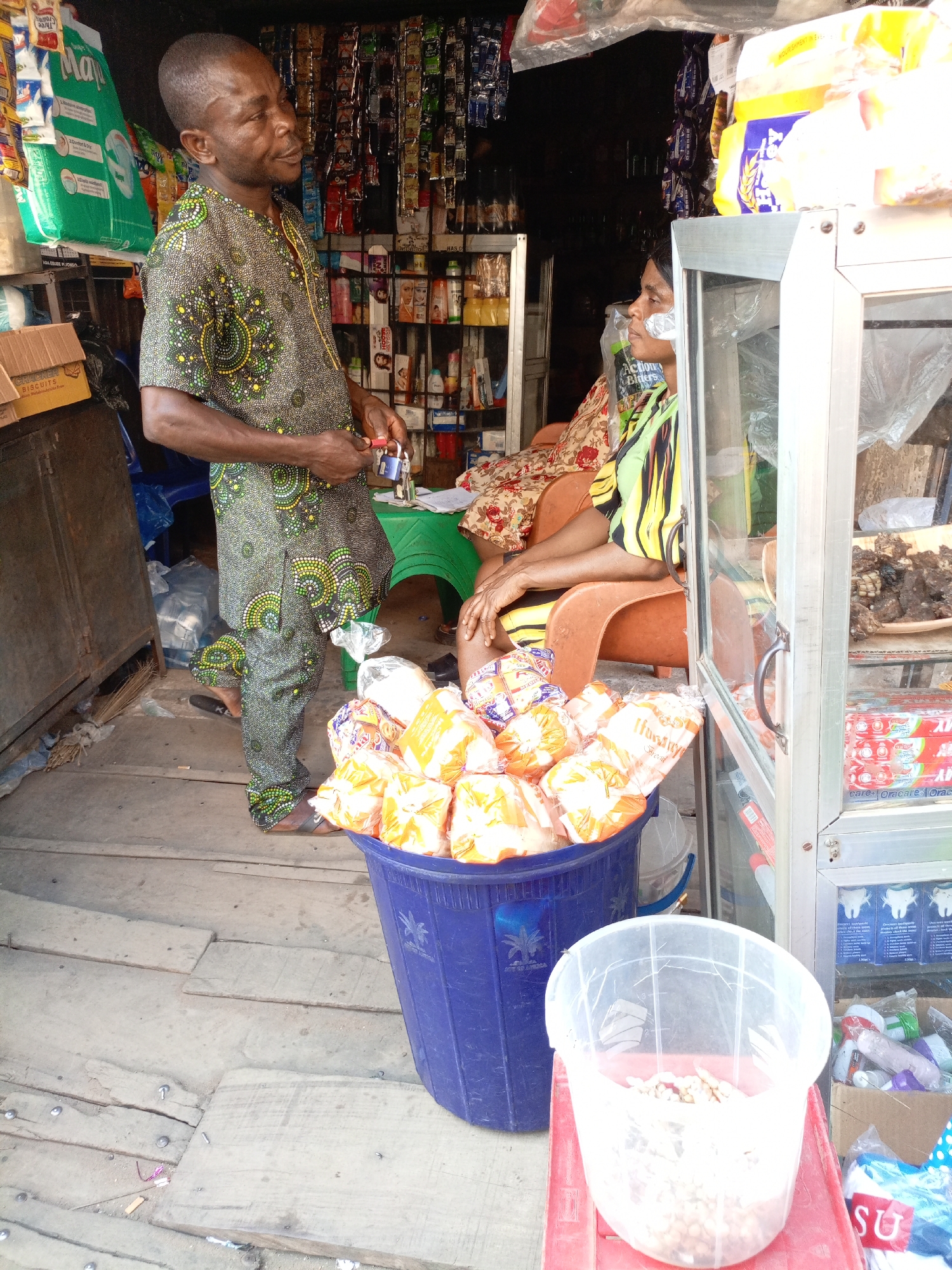 This image captures a vibrant scene inside a small shop or market stall, likely in Onitsha, Nigeria, during the daytime.
A man, with short dark hair and a light complexion, stands prominently in the center. He is wearing a patterned green and black short-sleeved top and matching trousers, typical of West African attire. In his right hand, he holds two small padlocks. He looks to his right, towards a seated woman or slightly past her. His expression is neutral, possibly engaged in conversation or observing the surroundings.
To the right, partially obscured by a metal-framed glass display case, a woman is seated on an orange plastic chair. She has dark skin and wears a multi-colored striped top over a floral-patterned garment. A piece of white tape or bandage is visible on her left cheek. She looks to her left, seemingly towards the man or into the shop.
The shop itself is densely packed with merchandise. In the background, shelves are filled with various packaged goods, including snacks, drinks, and toiletries, many hanging from hooks. A large green bag, possibly branded with "May", hangs on the left. On the left side, a cardboard box is visible, clearly labeled "BISCUITS".
In the foreground, a large blue plastic bucket is filled with numerous individually packaged items, likely bread or pastries, in clear plastic bags, some bearing the name "Hashir" and others "ASIAN". A palm tree logo is also visible on the bucket. On a red stool in front of the bucket, a clear plastic container holds small, light-colored nuts or seeds. The glass display case on the right contains more products, including boxes labeled "Oracare" (likely toothpaste) and a tray of what appears to be dried meat or fish on the top shelf. A green package near the woman reads "Action Bitters".
The floor is made of worn, uneven wooden planks. The overall atmosphere suggests a busy, informal retail setting.
