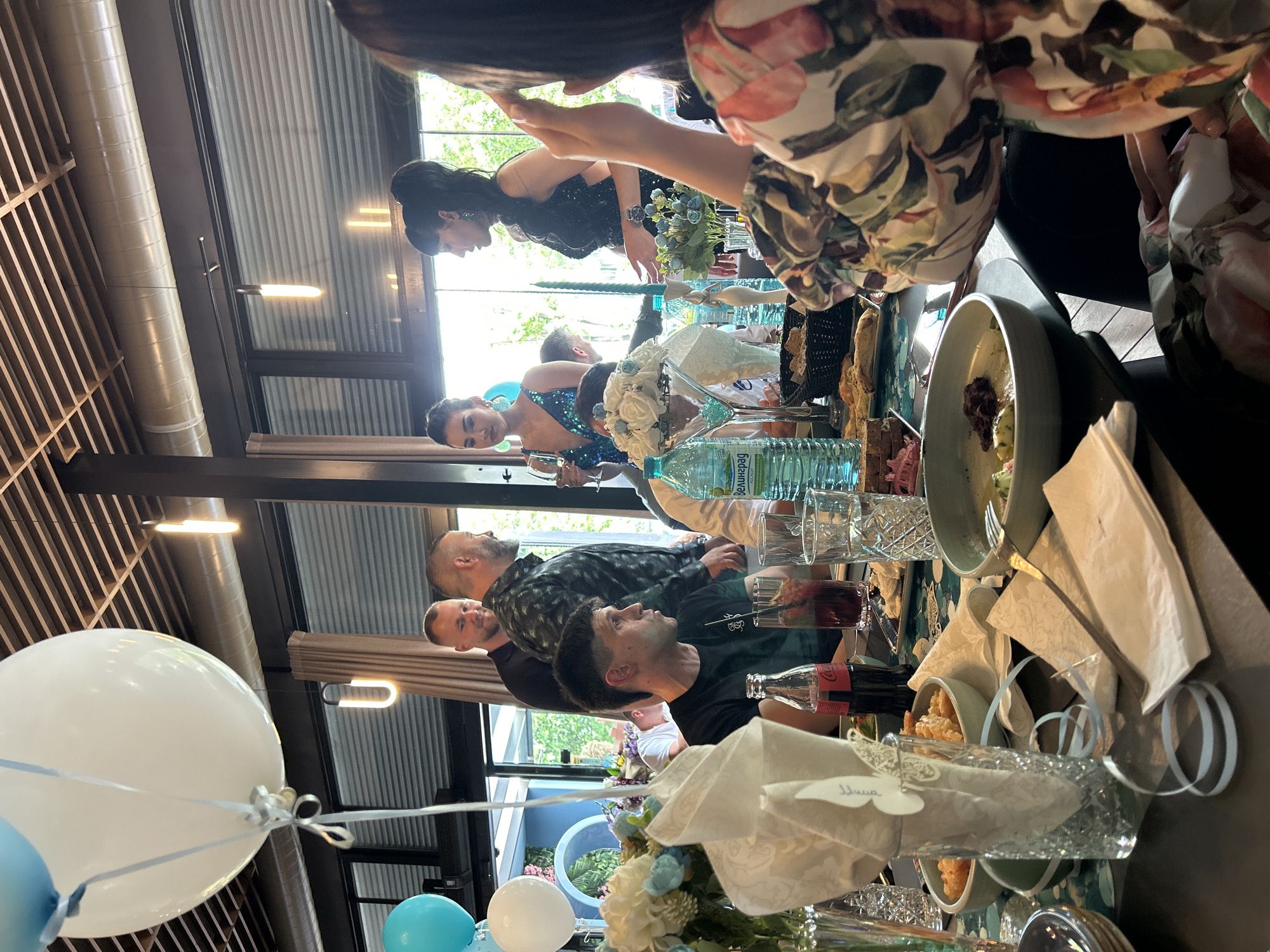 The image shows a group of people at a table, possibly at a celebration or event. There are at least two women in formal dresses, one in a blue sequined dress and another in a black sequined dress. Several men are also seated at the table. The table is set with glasses, bottles of water and Coca-Cola, plates, and decorative floral arrangements. Balloons are visible, suggesting a festive occasion. The location appears to be indoors, with large windows looking out onto greenery. The style of the interior, with its wooden beams and modern lighting, indicates a contemporary setting. It is difficult to ascertain if this is in Burgas, Bulgaria, based only on visual cues in the image.