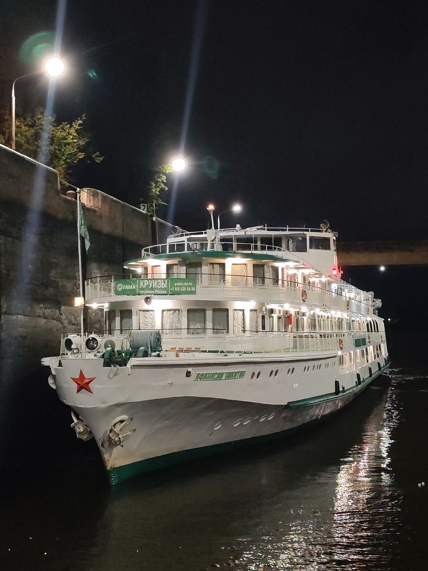 The image shows a multi-decked white passenger ship docked at night. The ship is illuminated by interior lights and has a red star emblem on its bow. A green banner with Russian text is visible on the side of the ship. Streetlights and trees are present on the left side of the image, casting light on a concrete wall. The ship is reflected in the dark water. The scene appears to be in a harbor or along a river.