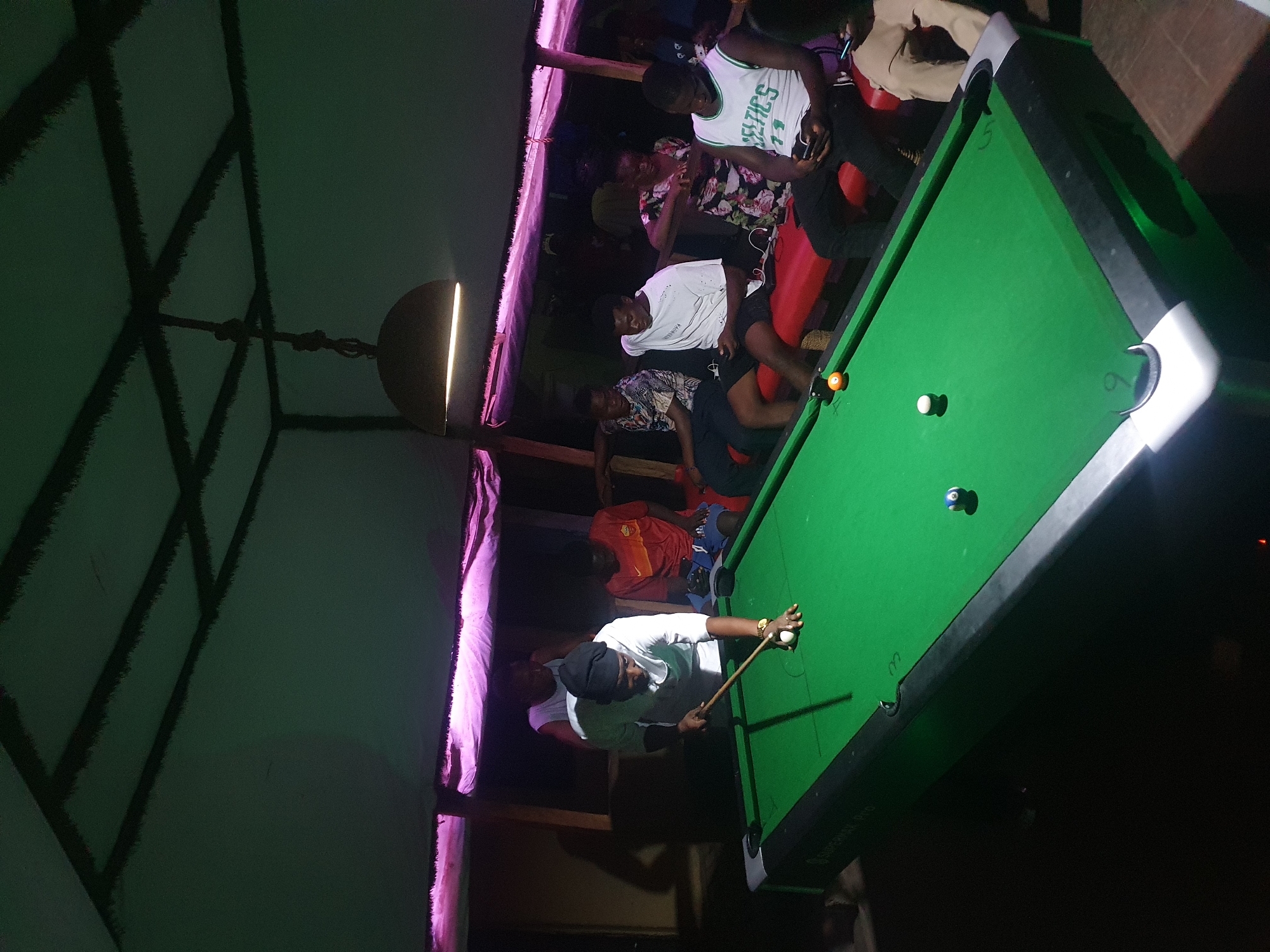 The image depicts an indoor scene at night in Lokoja, Nigeria, where a group of people are engaged in a game of pool. The central figure, wearing a white shirt and a dark head covering, is positioned to take a shot on a green-felt pool table, holding a cue stick. On the table, a white cue ball, an orange striped ball, and a blue solid ball are visible.
Several other individuals are gathered around the table, observing the game or socializing. One person wears a white Boston Celtics jersey with "CELTICS 14" prominently displayed. Another individual is seen in a floral patterned shirt, while a third wears a plain white t-shirt. Red seating is visible in the background.
The setting is dimly lit by artificial light sources, including a partially visible suspended light fixture and purple strip lighting running along a wall or beam. Above, a dark, crisscrossing structural framework, likely part of the ceiling, is silhouetted. The atmosphere appears relaxed and social, characteristic of a recreational venue. Faint text "SUPERPOOL PRO" is visible on the side of the pool table.