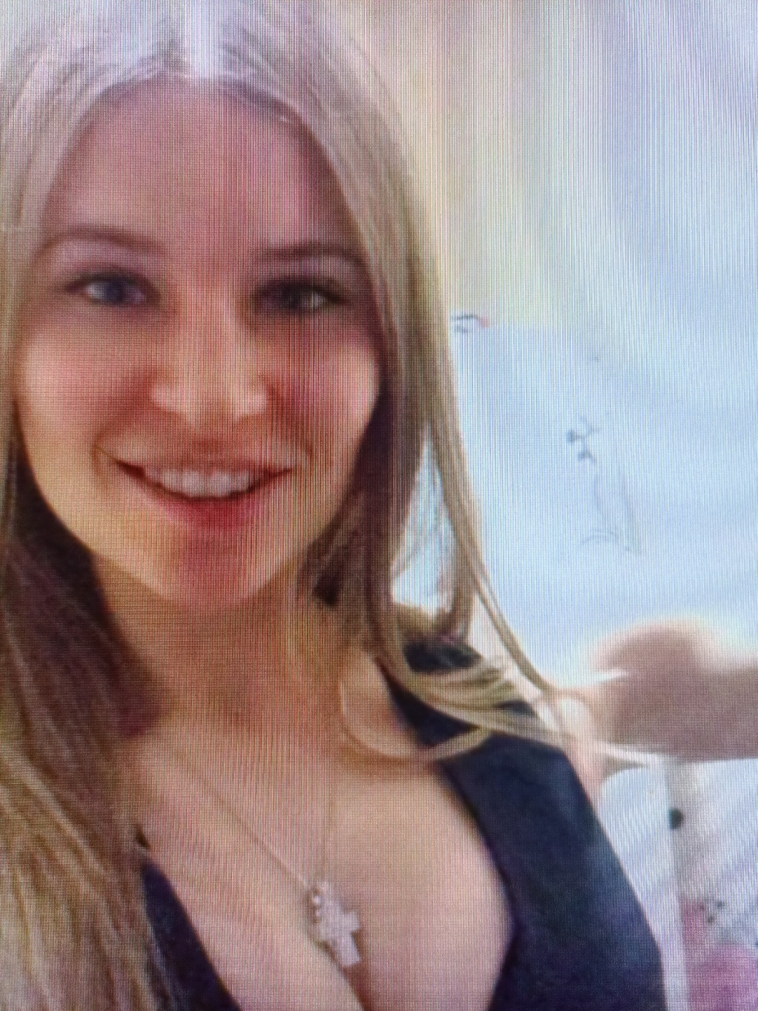 The image is a close-up, low-resolution photograph of a woman with blonde hair. She is smiling and wearing a dark top and a necklace with a cross pendant. The background appears to be a wall with some light-colored drawings on it. The photo quality suggests it was taken on a screen, possibly a television or computer monitor, as vertical lines are visible throughout the image. There are no discernible landmarks or features that would indicate a specific location within Nyagan, Russia.