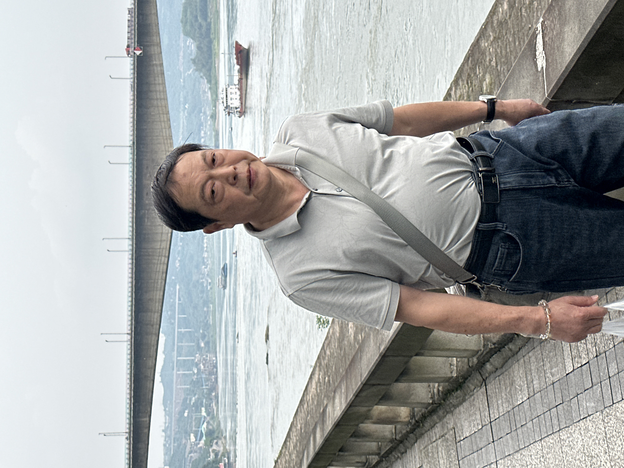 A man stands on a concrete ledge in Jiangjin, China. He wears a gray short-sleeved collared shirt, dark jeans with a black belt, and a watch on his left wrist, and a bracelet on his right. A gray strap crosses his torso. He looks towards the camera with a slight smile. Behind him is a body of water with a ship in the distance. To his left, a bridge structure spans the water. The sky is overcast. The ledge he stands on has a tiled wall beside it.