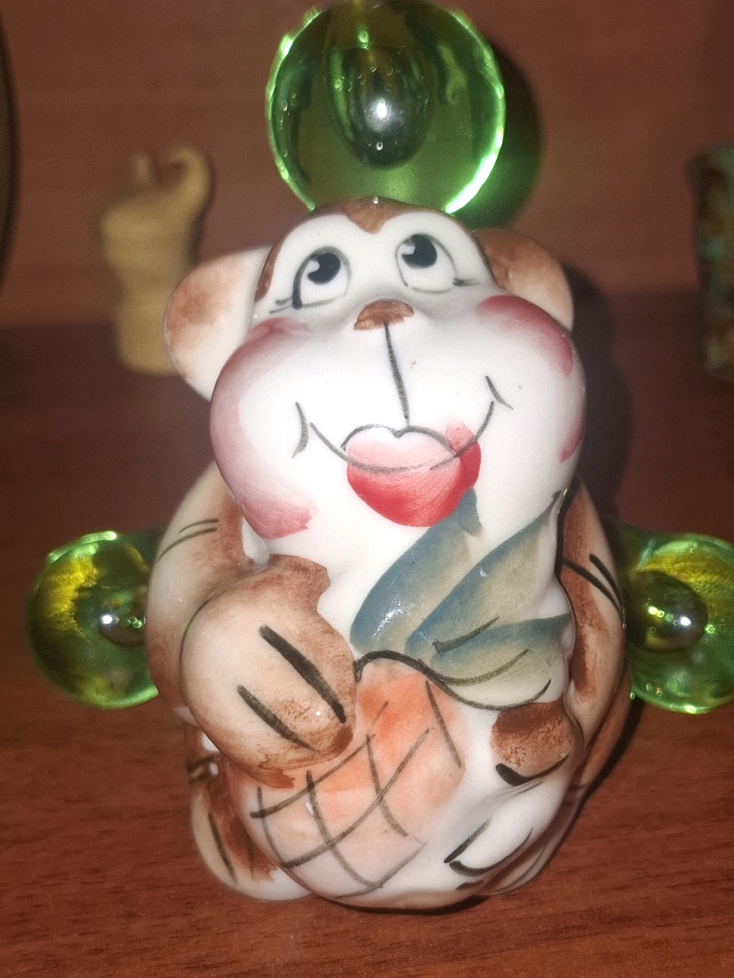 This is a close-up, eye-level, indoor photograph of a ceramic figurine of a monkey on a wooden surface. The figurine is white with brown and pink accents, and has large, expressive eyes and a cherry-like shape on its face. It is holding what appears to be an orange, segmented fruit, possibly a pineapple, and has green glass spheres attached to its sides and top. The background is a blurry pinkish-brown wall and hints of other objects are visible on the right and left. The scene is lit from above and to the front, creating reflections on the glass spheres. The location is not specified.