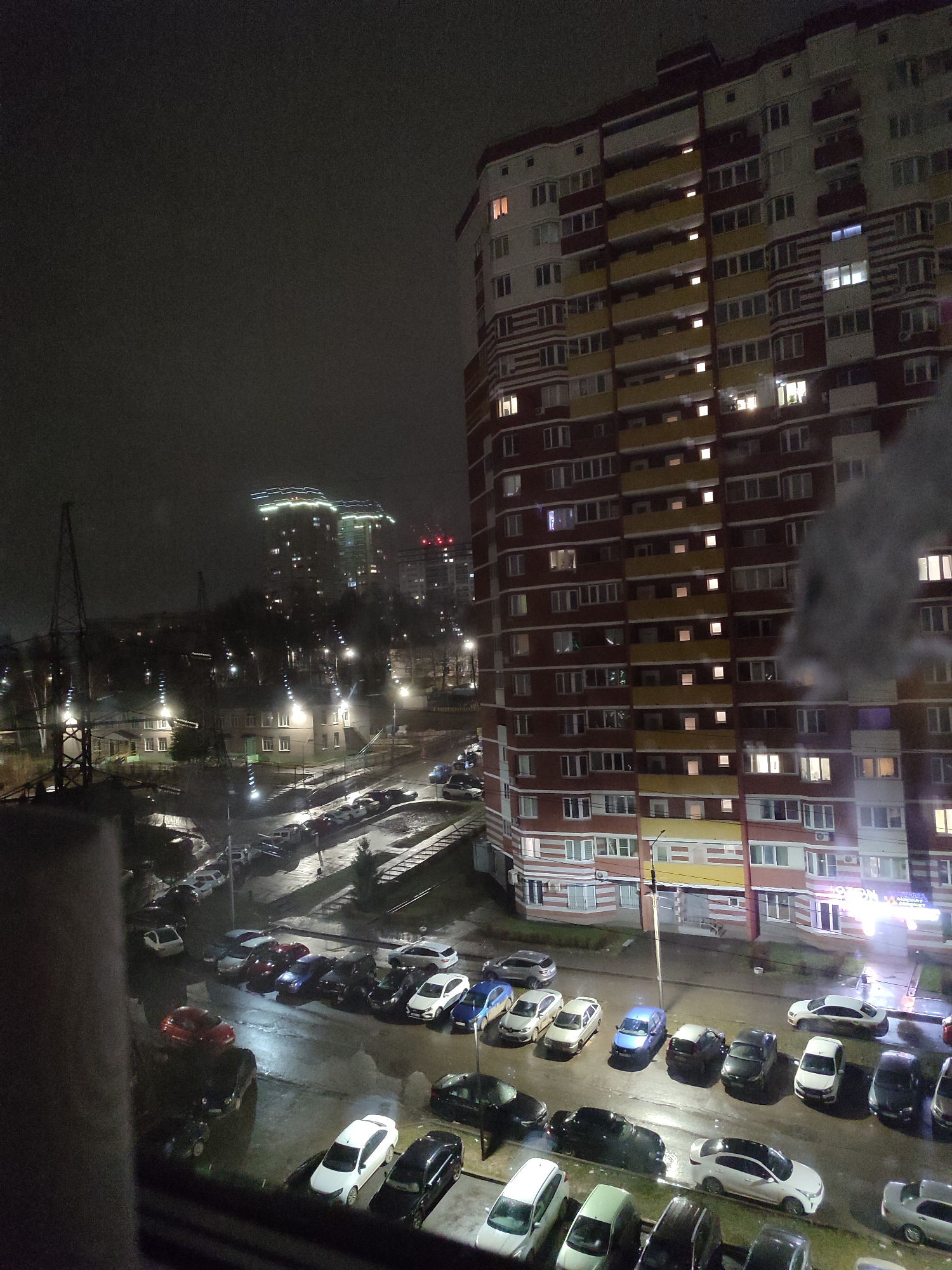 The image depicts a nighttime scene in Izhevsk, Russia, likely captured from an elevated perspective, possibly an apartment window. In the foreground and middle ground, a parking lot is visible, filled with numerous parked cars of various makes and models. The pavement appears wet, reflecting the ambient light and suggesting recent rain or damp conditions. Beyond the parking lot, there is a residential area featuring a high-rise apartment building with a mix of lit and unlit windows, indicating varying occupancy. Further in the background, more buildings are visible, illuminated and seemingly at a distance. Streetlights are scattered throughout the scene, contributing to the overall illumination of the urban environment. The sky is overcast, contributing to the somber and muted lighting conditions. Power lines and electrical towers are visible in the background, further indicating the urban setting.