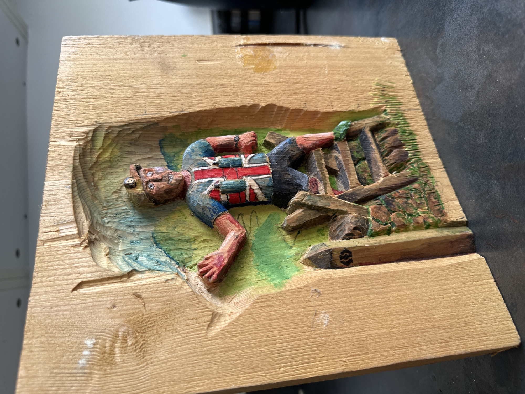 This is a detailed wood carving of a man wearing a Union Jack design on his shirt and shorts, climbing over a stile. He has a hard hat with a lamp on his head. The carving is mounted on a wooden block, with some of the background carved to depict a landscape with green and blue colors, and some grassy areas at the base. A small wooden post with a symbol on it is visible next to the stile. The carving is well-executed, with visible tool marks and painted details. The overall impression is that of a rustic and handcrafted piece. The man's stance suggests movement and effort as he navigates the stile. The Union Jack motif on his clothing suggests a British context. This carving is likely a decorative item, possibly representing a rambler or hiker in a rural British setting. Without further information, it is impossible to pinpoint its exact location in the city of Rugby, United Kingdom. However, the subject matter of climbing a stile and the Union Jack clothing suggest a connection to the English countryside, which is abundant in and around Rugby.