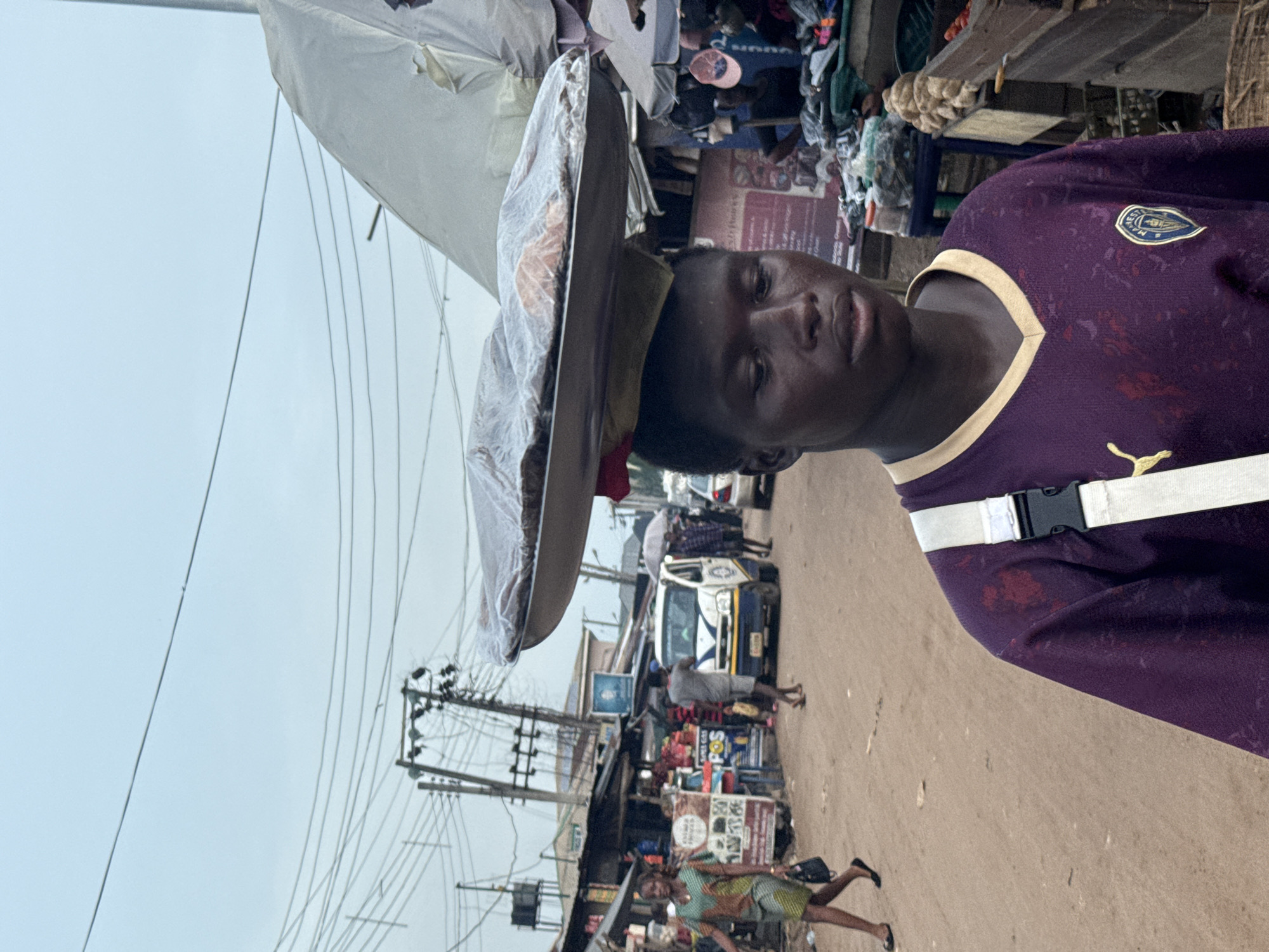 This image captures a vibrant street scene in Ihiagwa, Nigeria, under an overcast sky. The central subject is a young person, likely male, with dark skin, wearing a purple and beige sports jersey featuring a "Puma" logo and a crest (possibly a football club badge). He looks directly at the camera with a neutral expression. On his head, he expertly balances a large, flat object, possibly a tray or basket of goods, covered with a clear plastic sheet, suggesting he is a street vendor or hawker. A white strap with a black buckle is visible around his hip.
The background reveals a bustling outdoor market environment. The ground is unpaved and reddish-brown. Numerous market stalls and small shops line the street, displaying various items, including clothing. A dense network of utility poles and power lines crisscrosses the left side of the frame. In the mid-ground, a white and blue van or small bus is parked, surrounded by people. Another person in a green patterned dress is seen walking in the lower-left. Visible text includes the "Puma" logo on the jersey and partially discernible signs in the background like "Se Pos" and "PLAY." The overall scene depicts a lively day of commerce and movement.