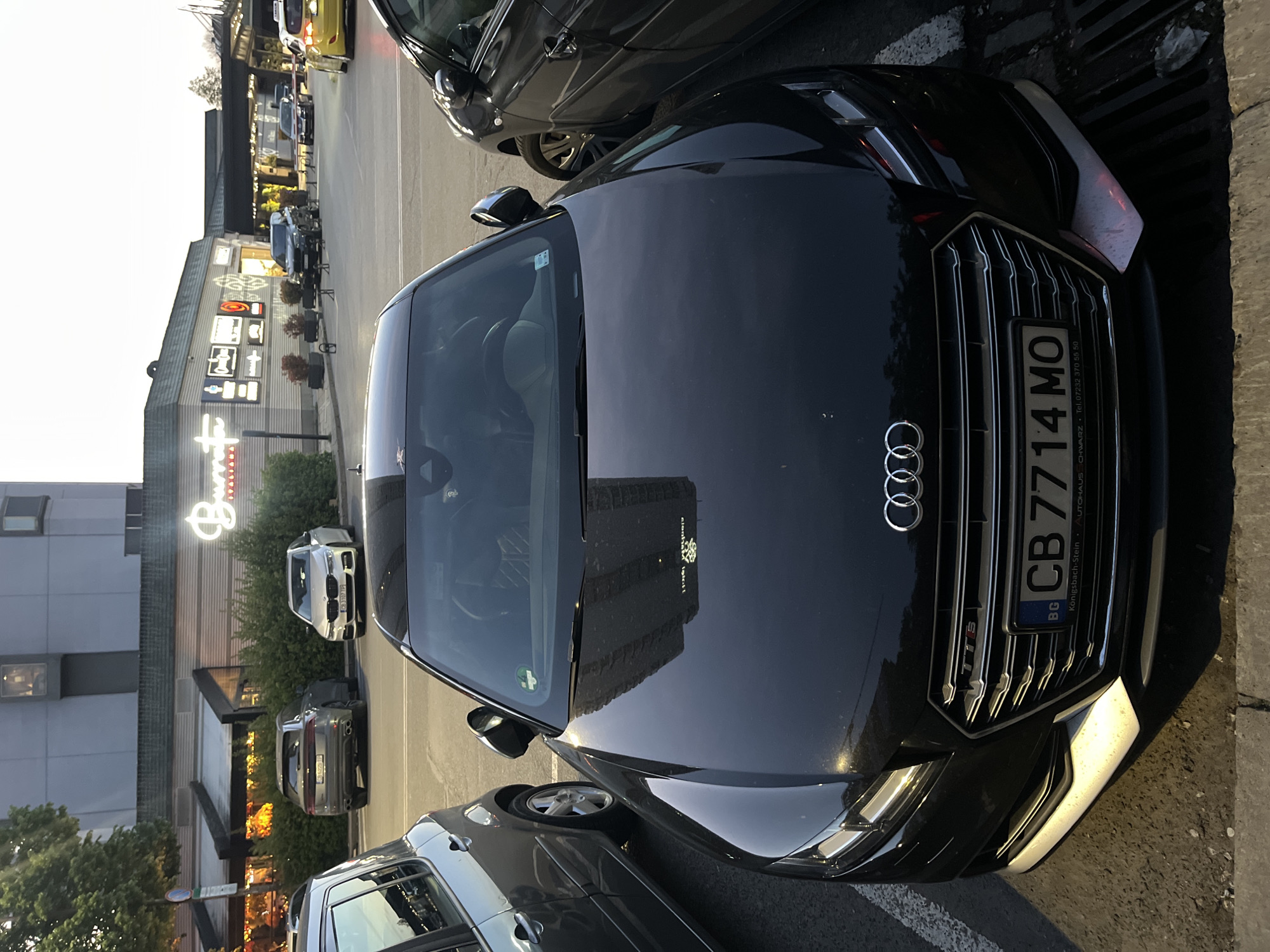The image depicts a black Audi automobile parked in a parking lot with a Bulgarian license plate number "CB 7714 MO". The reflection on the hood of the car suggests a building nearby, possibly located in Sofia, Bulgaria. In the background, there is a restaurant or commercial establishment with a lighted sign. Several other cars are parked in the vicinity. The image appears to be taken during twilight or evening. There are no people visible in the image.