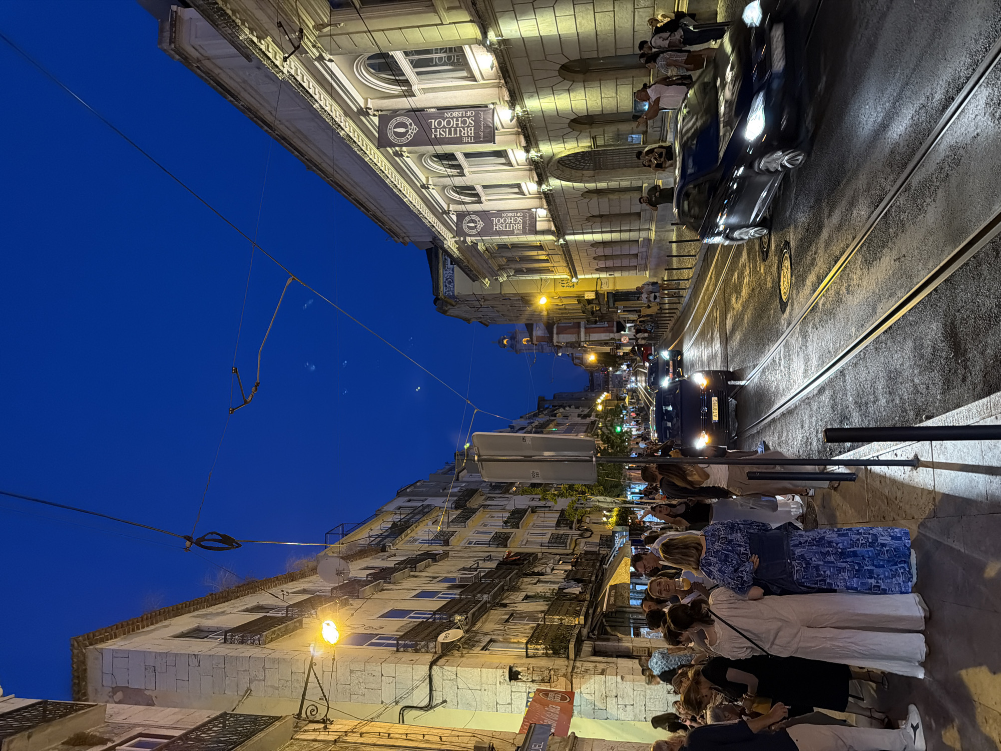 This is a low-angle, eye-level, wide shot of a street in Lisbon, Portugal, at dusk. The sky is a deep, clear blue. The street is lined with buildings on both sides, with a tram track running down the middle. On the left, a building with stone facade and balconies is illuminated by a street lamp. People are gathered on the sidewalk in front of shops and cafes. On the right, a building with classical architecture features arched windows and banners that read "THE BRITISH SCHOOL OF LISBON." Cars are driving on the street, their headlights illuminated. The scene is busy with pedestrian activity, suggesting a lively evening in the city.