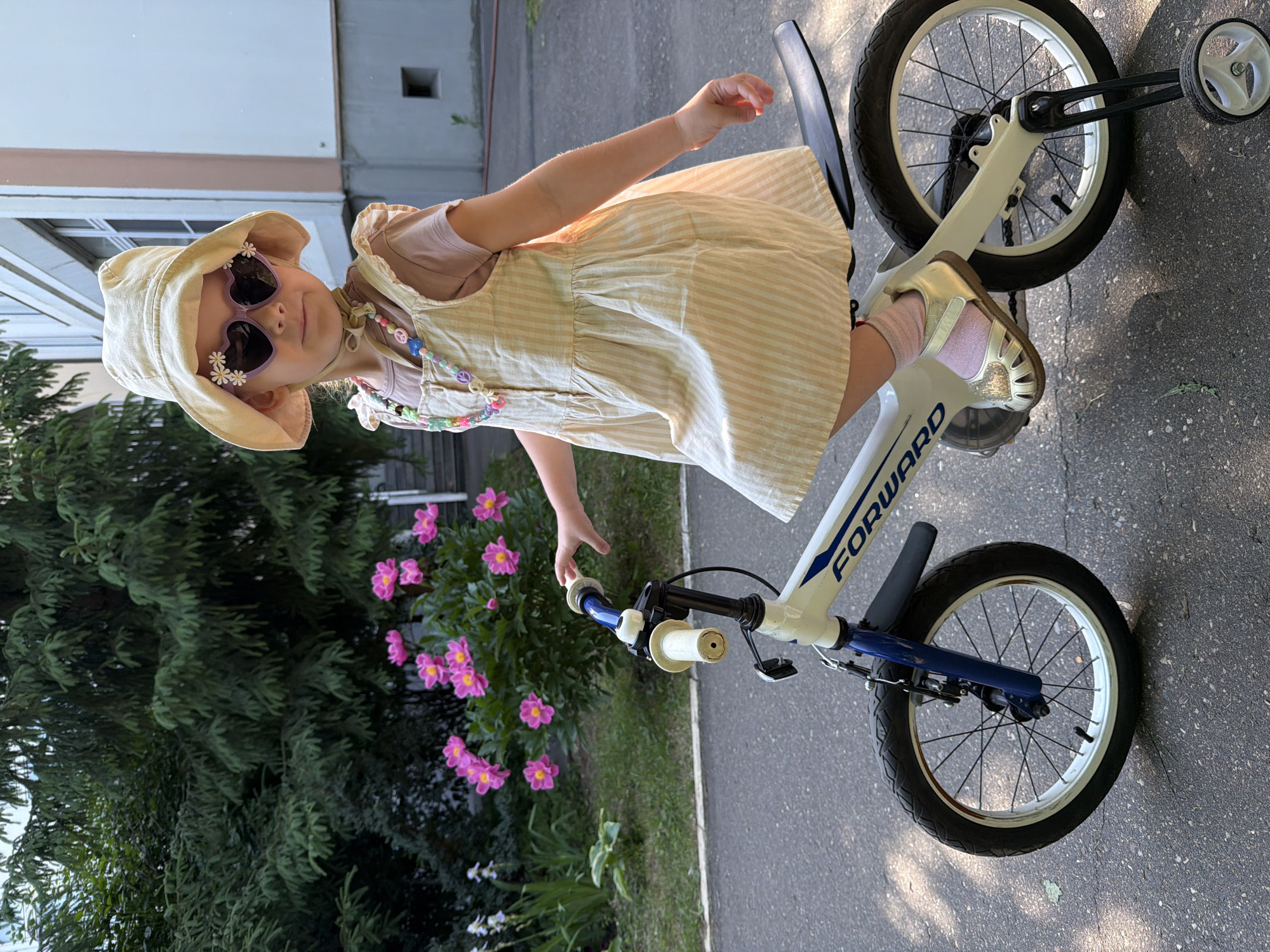 A young girl, wearing a light-colored bucket hat, heart-shaped sunglasses adorned with white flowers, a pink t-shirt, and a light-yellow and white striped pinafore dress, sits on a white and blue bicycle. She is also wearing a colorful beaded necklace with peace sign charms and pink socks with gold sandals. The bicycle has training wheels attached. The girl is positioned on an asphalt surface, likely a sidewalk or a path, with a grassy area to her left featuring pink peonies and other greenery. In the background, a building with light-colored walls and windows is visible, along with mature evergreen trees. The image is taken outdoors during daylight hours. The girl's left hand is extended towards the bicycle's handlebars. The scene appears to be in a residential or park area.