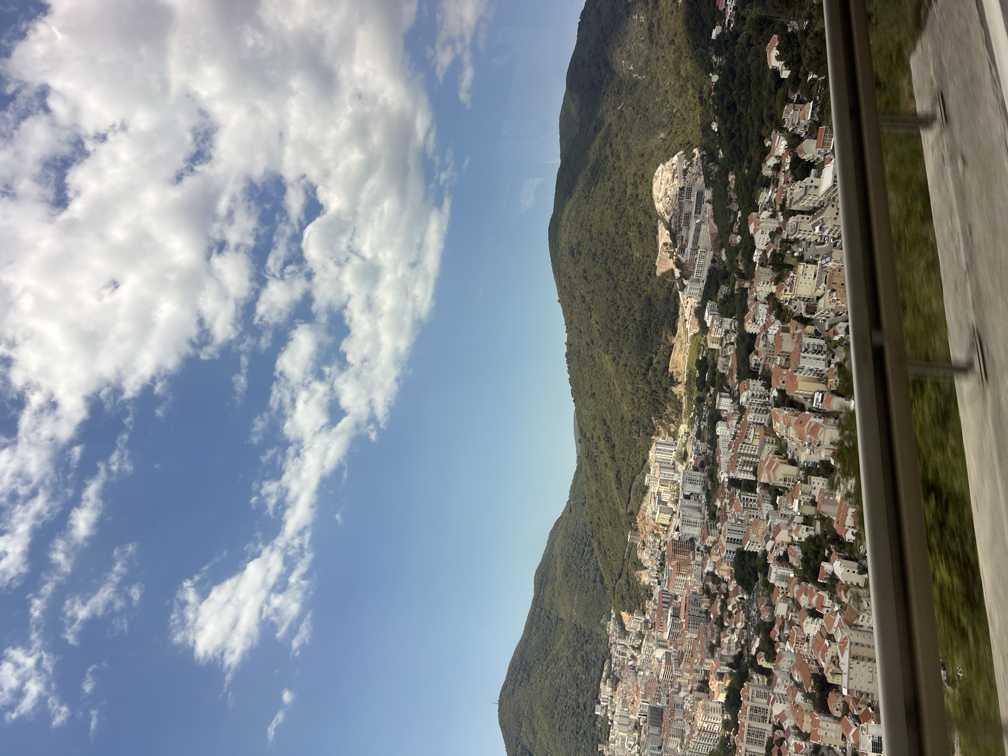A vertically oriented landscape photograph reveals a mountainous city scene under a partly cloudy sky, likely taken from an elevated or moving vantage point, possibly a cable car or high building.
To the left, a blue sky is punctuated by fluffy white clouds. Occupying the center of the frame is a densely forested mountain range, its dark green foliage contrasting with the lighter hues of the sky. At the foot of the mountains lies a dense cluster of buildings, characterized by their red-tiled roofs and white or light-colored facades. Some buildings appear to be under construction on the mountainside, suggesting ongoing development.
The right edge of the frame displays part of a window frame or barrier, with a vertical metal support and reflective glass, indicating the perspective from within a structure.
The city is likely Budva, Montenegro.