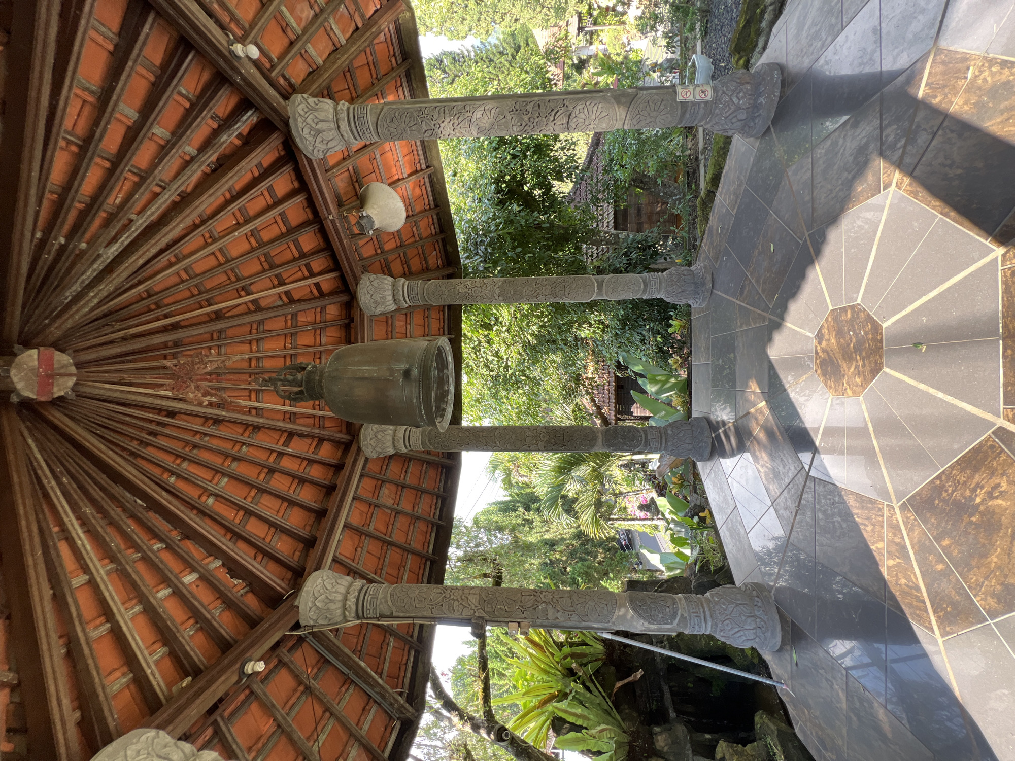 The image shows the interior of a gazebo in Pupuan, Indonesia. The gazebo has a tiled roof supported by four carved stone pillars. A large bell hangs from the center of the roof. The floor is tiled with a geometric pattern of dark and brown tiles. Lush green vegetation and trees surround the gazebo, and a small building is visible in the background. The scene is bathed in sunlight, creating strong shadows on the floor.