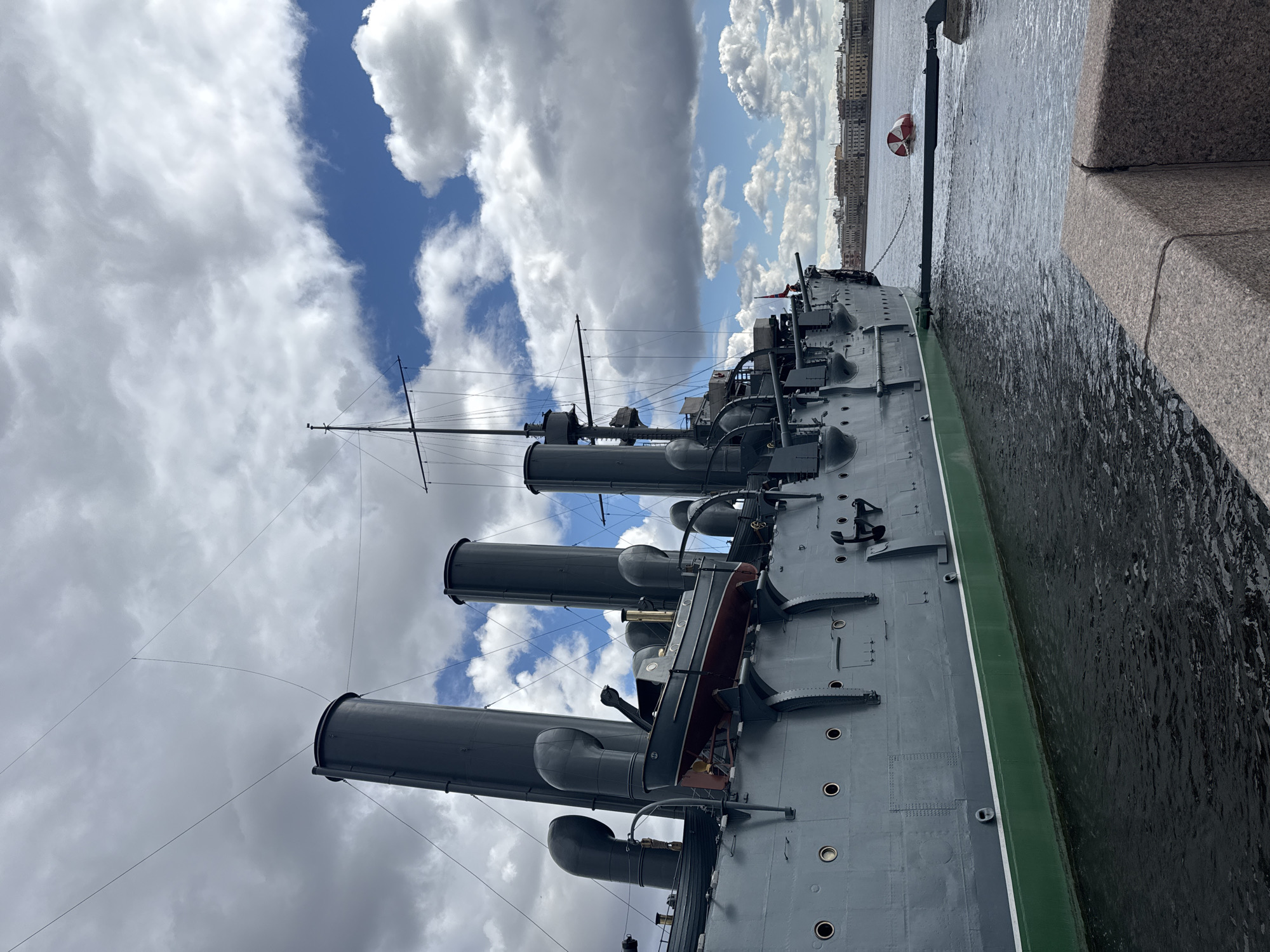 The image shows the Cruiser Aurora, a Russian protected cruiser, permanently docked in Saint Petersburg, Russia. The vessel is painted in a gray color scheme with a green and white stripe at the waterline. Three prominent smoke stacks are visible, along with a lifeboat mounted on the side of the ship. Masts and rigging extend upwards towards a cloudy sky with patches of blue. The ship is moored alongside a granite embankment, with water reflecting light, and building visible across the water in the distance. A red and white buoy is visible near the ship.