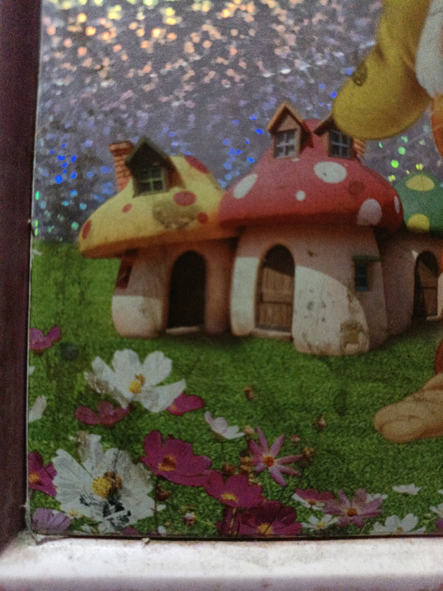 The image shows a close-up of a sticker or printed material featuring a whimsical, cartoonish scene. In the foreground, there is a green grassy area with various flowers, some white with yellow centers, and others pink. The flowers appear to be stylized representations. Behind the flowers are two mushroom-shaped houses. The house on the left has a yellow cap with red spots and a light-colored base with a dark arched doorway. The house on the right has a red cap with white spots and a similar light-colored base, also with a dark arched doorway and a small square window. The background has a sparkly, iridescent texture with hints of blue, purple, and pink lights, suggesting a starry or magical sky. The overall image has a grainy, low-resolution appearance. There is no discernible connection to West Jakarta, Indonesia.