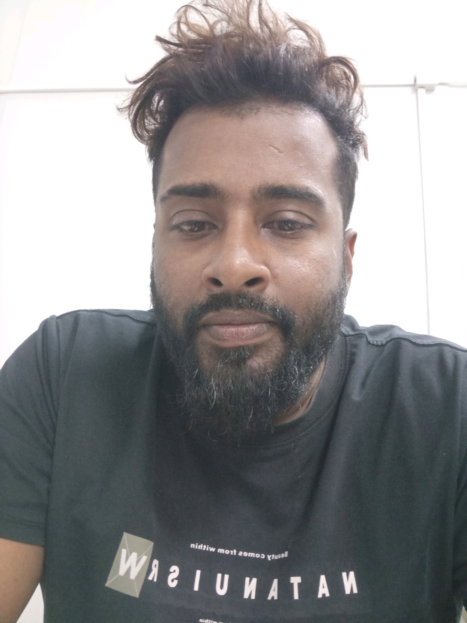A head and shoulders, close-up shot of a man with dark skin, a full beard, and styled, dark brown hair facing the camera. He is wearing a dark gray t-shirt with a graphic design and text. The background is a plain, white wall with what appears to be vertical paneling. The man's expression is neutral. The image was taken in the city of Kiryat Gat, Israel.