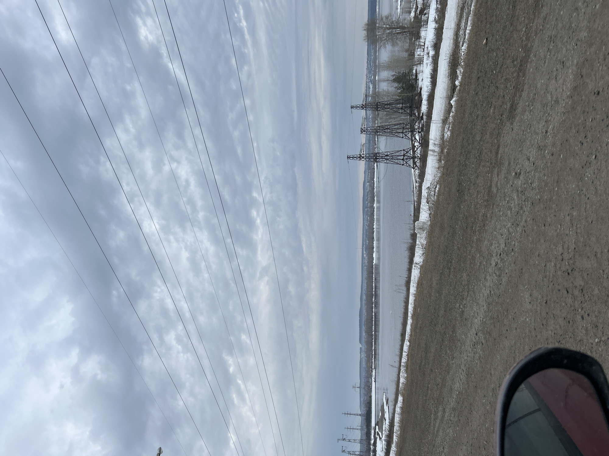 The image depicts a landscape in what appears to be the city of Chaikovsky, Russia, based on the presence of power lines and a body of water. The sky is overcast with a thick layer of clouds. Power lines stretch across the sky, converging towards the horizon. In the midground, a body of water, possibly the Kama River, is visible. Power transmission towers line the edge of the river, some covered with snow. Patches of snow are also visible along the shoreline, indicating a cold climate. The foreground is dominated by an unpaved road or area of gravel, with the side mirror of a vehicle visible in the bottom right corner, suggesting the photo was taken from a moving car. The image lacks people, objects, actions and interactions.