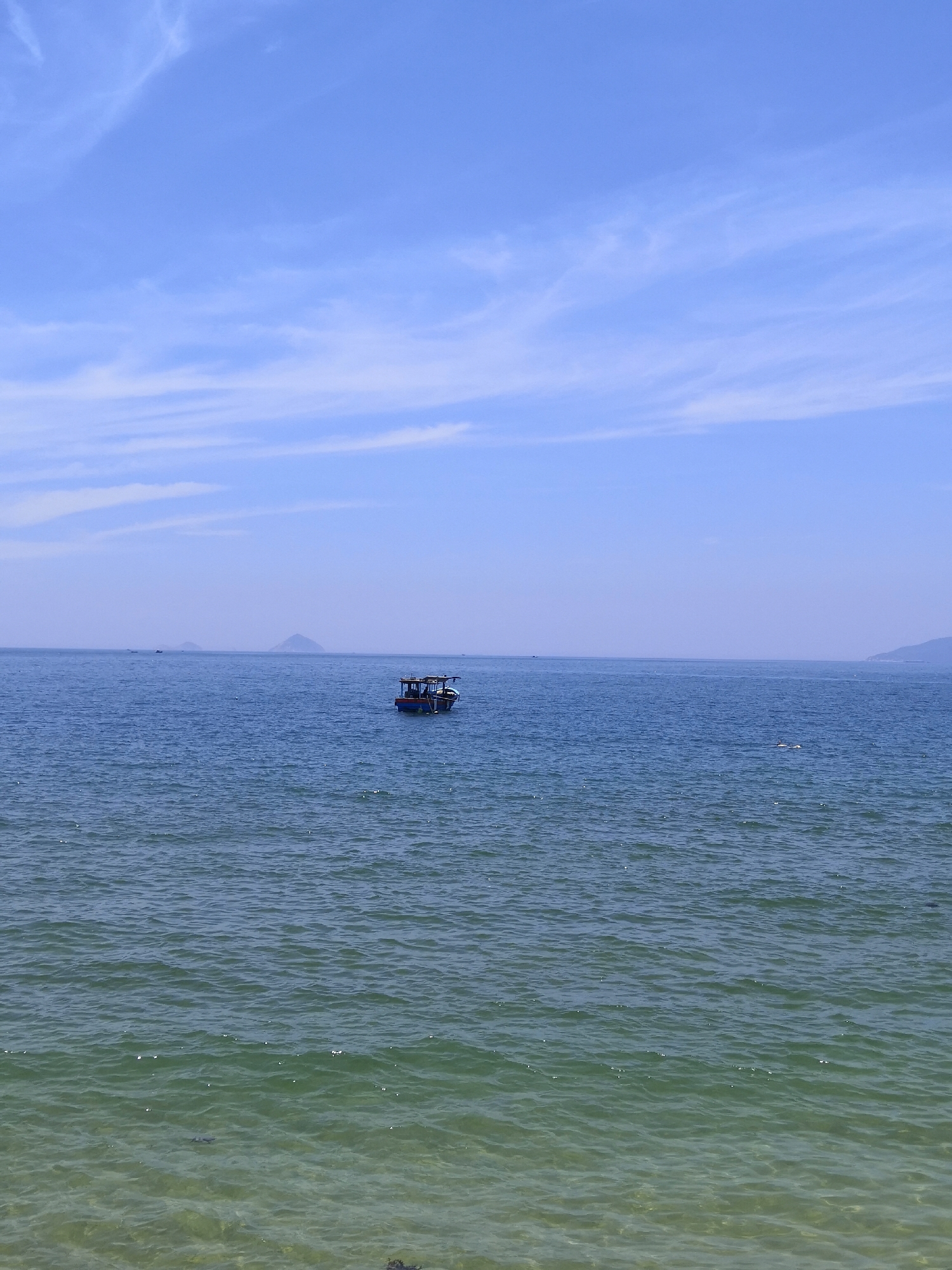 This image depicts a coastal scene in Bac Nha Trang Ward, Vietnam, on a clear day. The upper two-fifths of the frame are dominated by a vast, light blue sky featuring scattered, thin, white cirrus clouds, particularly in the upper left and horizontally across the mid-sky. The horizon line is flat and distinct.
Below the horizon, the sea extends into the distance, appearing a deep cerulean blue. Several indistinct, darker landmasses, possibly islands or distant hills, are visible on the horizon in the central and far-right portions of the frame. A single, small to medium-sized motorboat, characterized by a dark hull and a lighter, possibly white or light blue, cabin or canopy structure, is positioned in the mid-right area of the water. The boat appears stationary or adrift.
The water surface is covered with small, consistent ripples, reflecting sunlight as numerous tiny white specks. As the water approaches the foreground, its color transitions to a clearer, greenish hue, indicating shallower depths. The transparency of the water in the immediate foreground allows glimpses of the submerged seabed, where darker, irregular shapes, consistent with rocks or aquatic vegetation, are visible at the very bottom edge of the frame. No individuals are discernibly present on the boat or in the water.