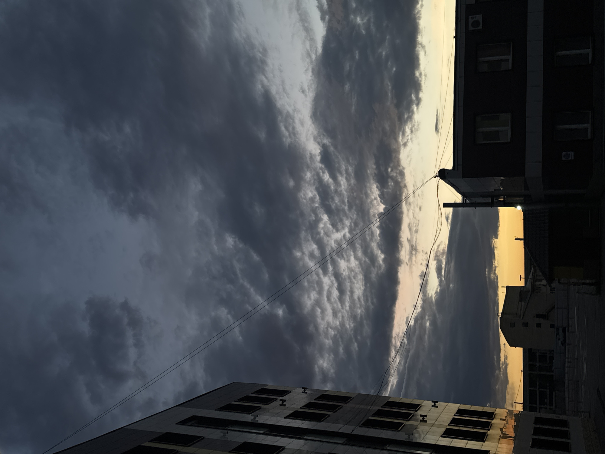 This image depicts an atmospheric urban landscape, likely captured at either sunrise or sunset in Perm, Russia. The sky is a dominant feature, filled with heavy, dark grey clouds that stretch across much of the frame. There are visible lighter, almost white, patches within the cloud cover, suggesting varying thickness or breaks. Towards the right side, a strip of vibrant golden-yellow light is visible near the horizon, contrasting sharply with the somber clouds and indicating the sun's low position.
Several buildings are silhouetted against the dramatic sky. On the right, a multi-story building with a dark facade, possibly an apartment or office block, is visible. It features several rectangular windows, some appearing illuminated or reflective, and at least two external air conditioning units. Thin power lines extend from this building across the sky, adding an urban element. In the foreground on the left, another modern-looking building with a reflective, dark glass facade is partially visible, also showing numerous large, dark rectangular windows. The reflections on its surface subtly mirror the sky's light.
The scene conveys a quiet, dramatic mood, characterized by the interplay of light and shadow and the contrast between the expansive, cloudy sky and the city structures. No people or active events are visible. The image emphasizes the architectural forms within an urban setting under striking atmospheric conditions. There is no discernible text within the image.