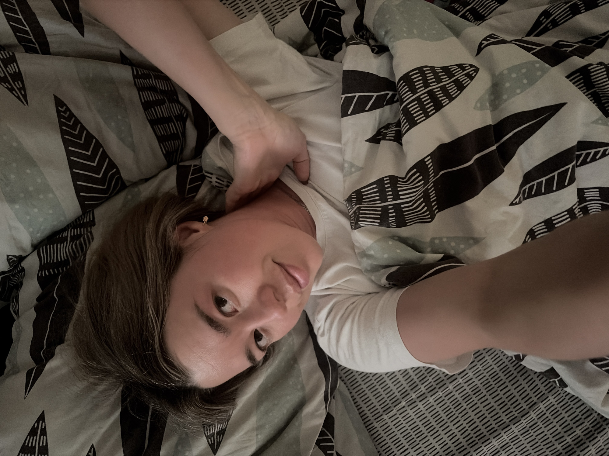 A woman lies in bed, partially covered by a patterned duvet. The duvet has a pattern of stylized leaves in black and grey on a light background. She is wearing a white t-shirt. Her left arm is bent with her hand near her neck, slightly pulling at the collar of her t-shirt. Her right arm is extended towards the camera, suggesting she is taking a selfie. She has short brown hair and is wearing an earring. The location is unspecified, but the prompt mentions the city of Chaikovsky, Russia, which might be relevant.