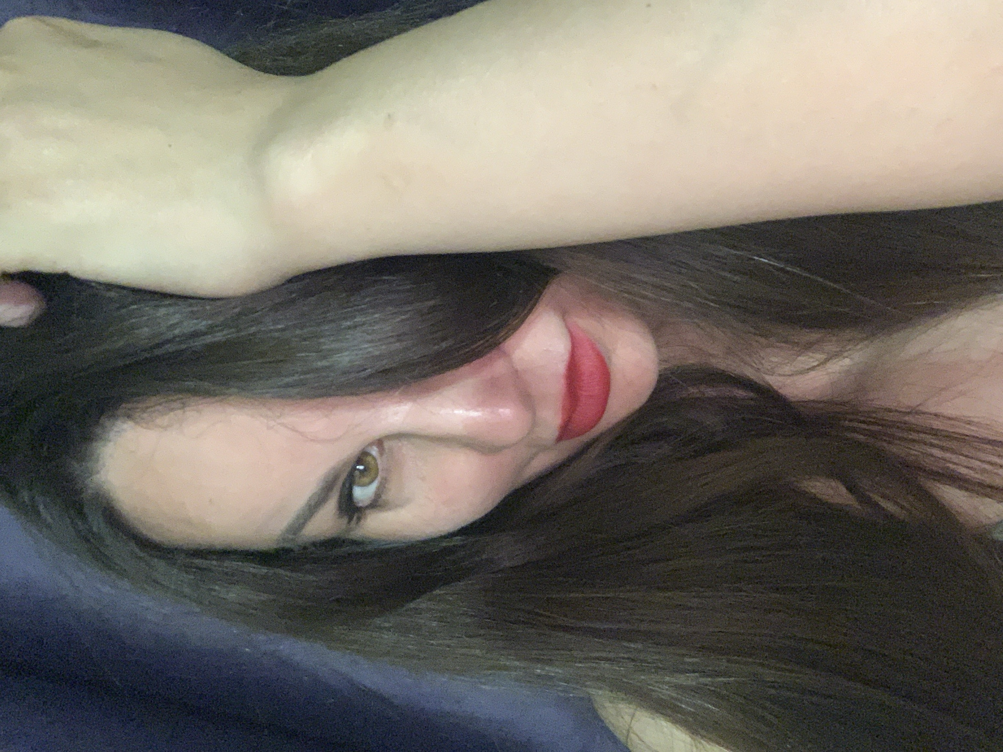 The image is a close-up, slightly angled portrait of a woman. She has dark hair and fair skin. She is wearing red lipstick. Her eyes are brown. Her left arm is raised over her head and partly obscures her face. The background is a dark, blurry texture. There is no city visible in the image.