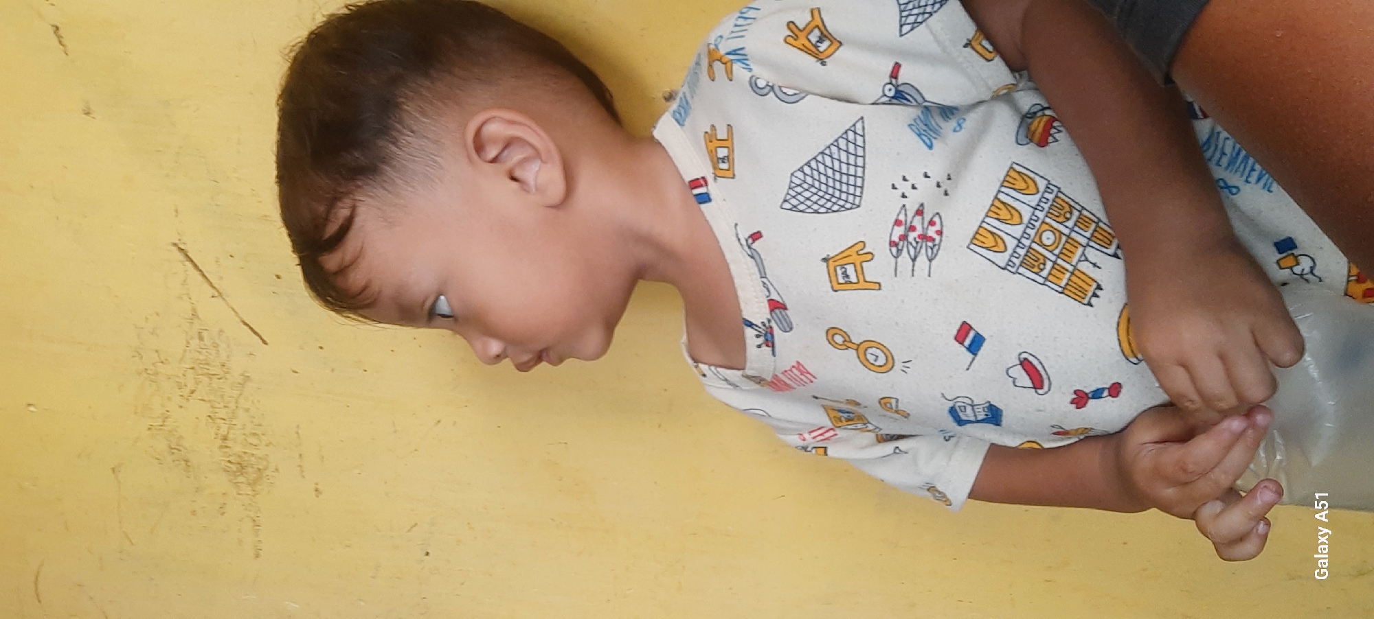 A young child, viewed from the side and slightly from above, gazes towards the left. The child has short, dark hair, styled with a fade, and light brown skin. They are wearing a short-sleeved t-shirt with a repeating pattern of various cartoon-like illustrations, including a white hat with a red band, what appears to be a bread loaf, and various abstract shapes and objects. The child's hands are visible in the foreground, one holding a clear plastic bag. The background is a textured, yellow wall. The image contains a watermark in the bottom left corner that reads "Galaxy A51". The location of Selopuro, Indonesia is not evident from the visual information provided in the image.