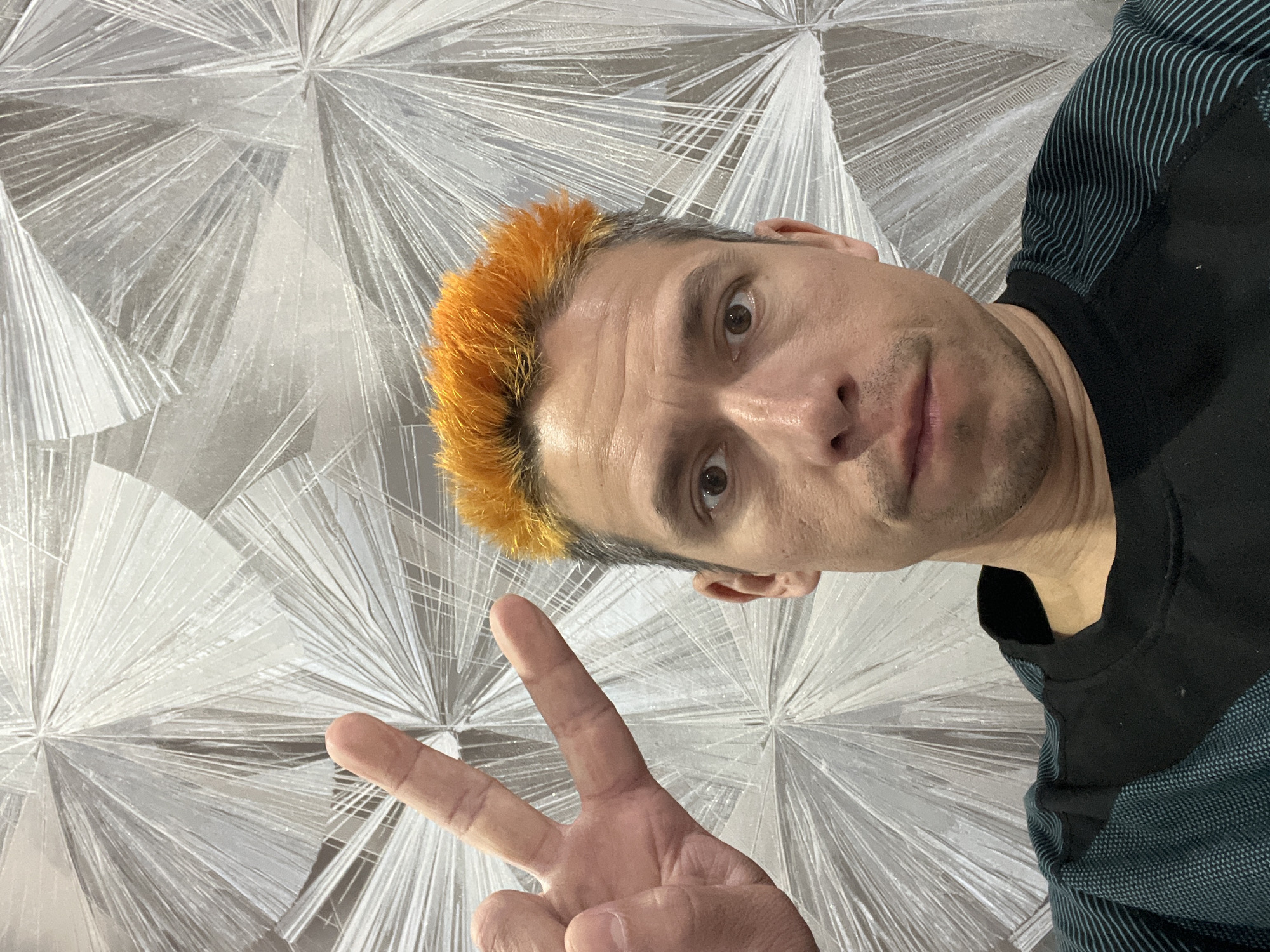 A man with orange hair poses indoors, making a "peace" sign with his fingers. He is wearing a dark shirt and the background is a silver, starburst-patterned wall. While the location is said to be in the city of Chaikovsky, Russia, the photograph itself does not provide any visual information to confirm this.