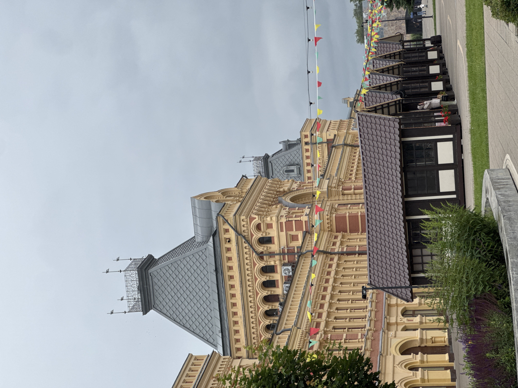 The image captures a section of Nizhny Novgorod's historic Red Market (Krasnaya ploshchad) on an overcast day. The primary structure in the background is the grand building of the market itself, characterized by its striking yellow facade with red brick detailing and ornate architectural elements, including arched windows and decorative cornices. A prominent grey, mansard-style roof with a decorative tower and weather vanes tops the structure. In the foreground, a row of small, dark-colored market stalls with brown tiled roofs are lined up. These stalls have large windows and doorways, and one person is visible standing near an entrance. Festive colorful flags are strung across the scene, both above the market stalls and along the facade of the main building, suggesting a public event or festive atmosphere. Greenery, including trees and bushes, is visible on the left side and in the foreground, alongside a concrete planter. The ground is paved with cobblestones or interlocking pavers. The overall environment appears to be a public square or pedestrian area.