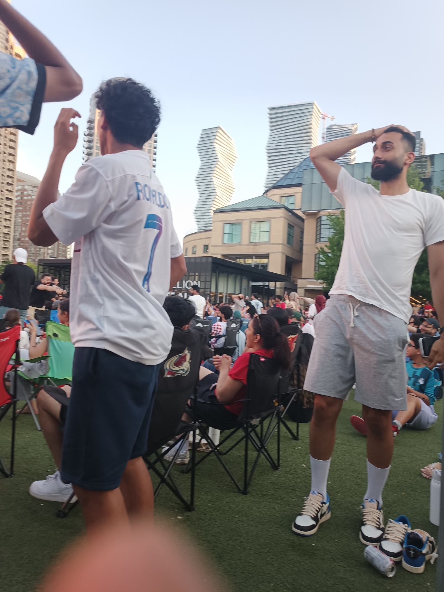 The image depicts an outdoor gathering of people, possibly attending an event, with a backdrop of modern architecture. In the foreground, two young men are the primary subjects. The man on the left, facing away from the camera, wears a white t-shirt with the word "RORDOS" and the number "7" emblazoned on the back, paired with dark blue shorts. He is seated on a folding chair. To his right, another man, facing the side, wears a white t-shirt and light gray shorts. He has his hand on his head, looking towards the right. Both men are wearing athletic footwear.
The environment is a green, artificial turf surface where numerous people are seated in folding chairs, some holding phones. Behind the individuals, a series of tall, distinctive buildings with rippled facades rise against a clear sky, characteristic of the Mississauga city skyline, particularly the area around Square One. Structures like the Mississauga City Hall and the City Centre are visible, suggesting the location as Mississauga, Ontario, Canada. The overall atmosphere appears to be casual and social, with people enjoying an outdoor event.