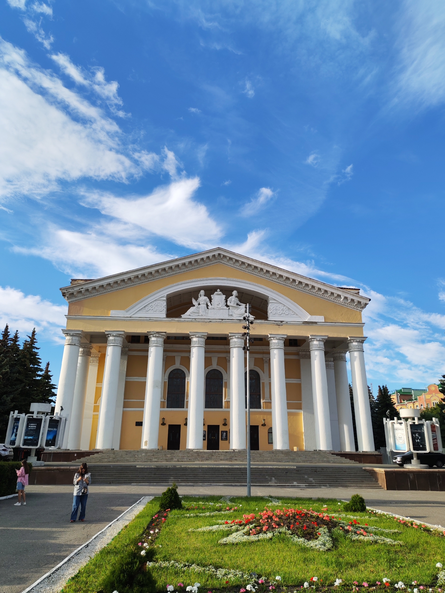 The image depicts a wide, low-angle shot of a grand building with neoclassical architecture, likely a theater or government building, under a bright blue sky with scattered white clouds. The building is painted a soft yellow and features a prominent portico supported by large, white Corinthian columns. Above the columns, a pediment contains sculptures, including figures of women. The building has arched windows and a grand staircase leading to its entrance.
In front of the building, a well-maintained lawn is decorated with a flowerbed featuring a variety of colorful flowers, predominantly red and white. Two women are visible in the foreground, one standing and looking at her phone, and another further to the left walking away from the camera, both dressed casually. Behind them, to the left, are tall evergreen trees and a display structure with screens showing images and text. To the right, another similar display structure is visible with a car parked nearby.
The scene appears to be set during the daytime, with the sunlight casting shadows and illuminating the building and its surroundings. The weather seems pleasant, with a clear blue sky and dynamic clouds suggesting a partly cloudy day. The presence of people suggests it's an accessible public space, and the landscaping indicates a civic area, possibly a plaza or park.
The text on the display structures is partially visible. On the left display, one screen shows text including "ТЕАТР", "АФИША", and "СЕЗОН". On the right display, text visible includes "СИМФОНИЯ", "КОНЦЕРТ", and "КЛАССИКА". These indicate that the building is likely a theater, and the displays are advertising events. The location context provided is Yoshkar-Ola, Russia.